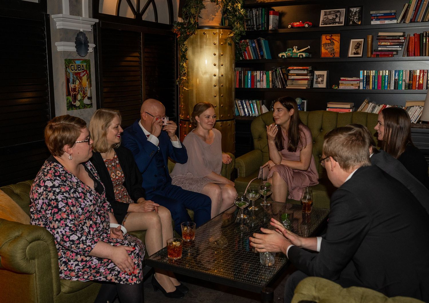 Wedding guests seated in cozy lounge area with bookshelves chatting at intimate reception gathering.