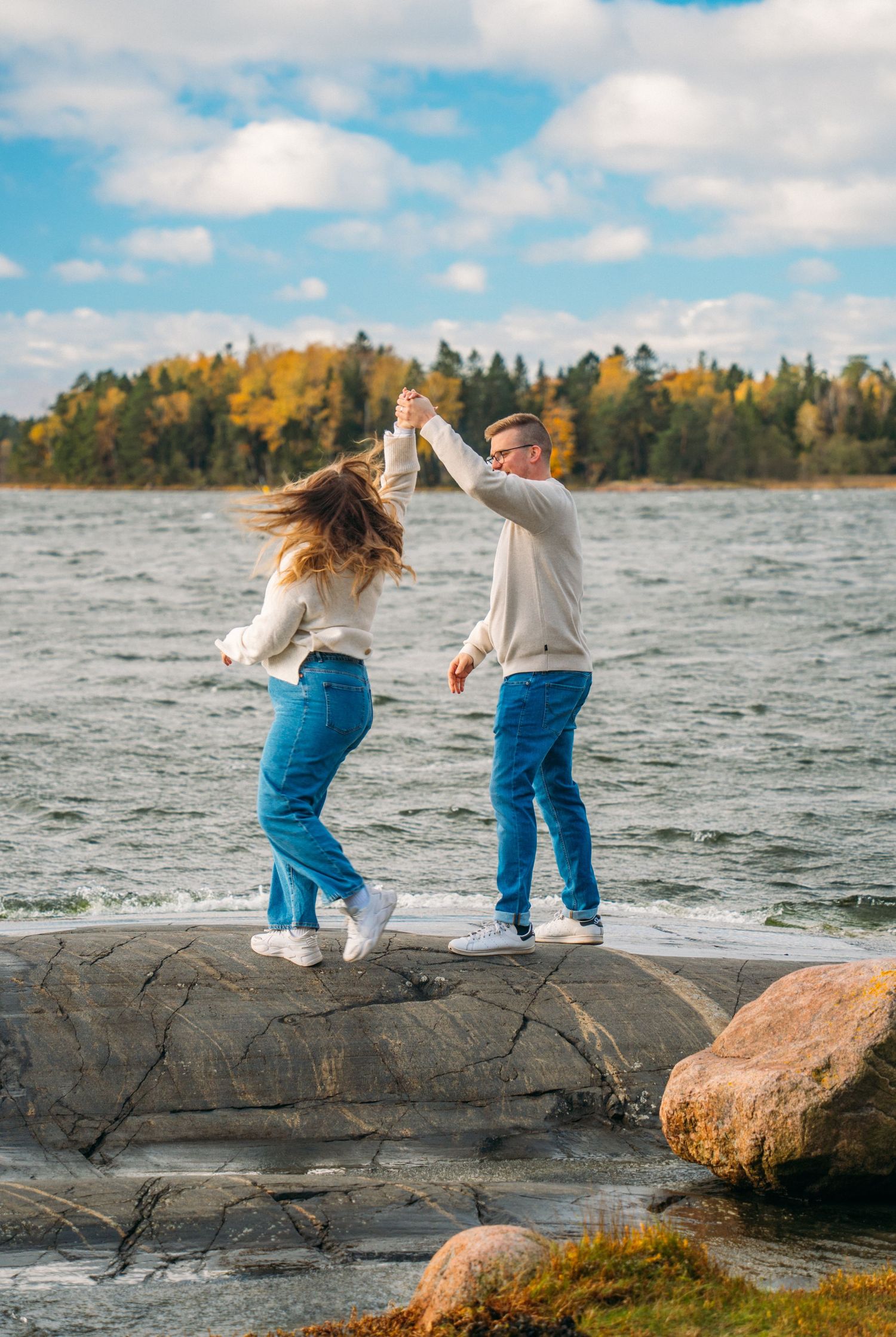 A couple dances joyfully on rocky shoreline, holding hands in matching blue jeans and white sweaters with autumn trees behind.