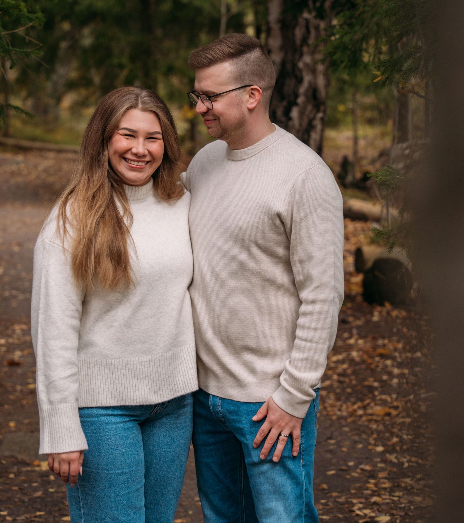 A smiling couple in matching beige sweaters and blue jeans stands together on a forest path in autumn.