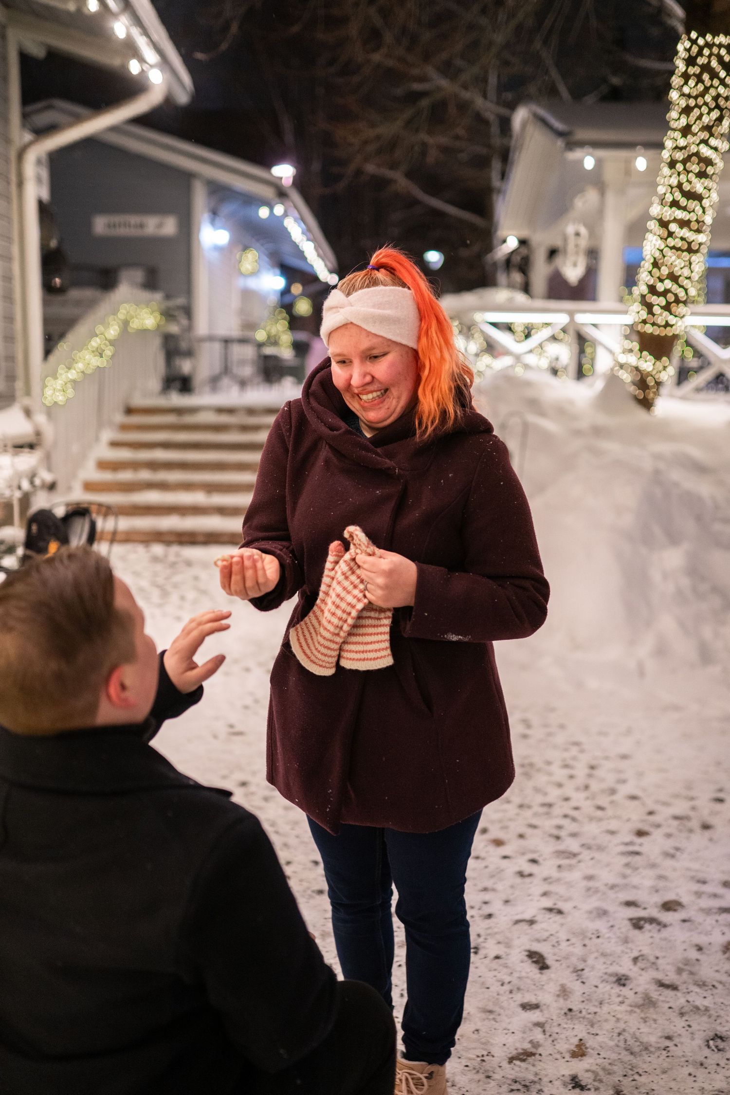 Woman with orange hair in winter coat smiles outdoors at night, snow-covered ground and Christmas lights in background.