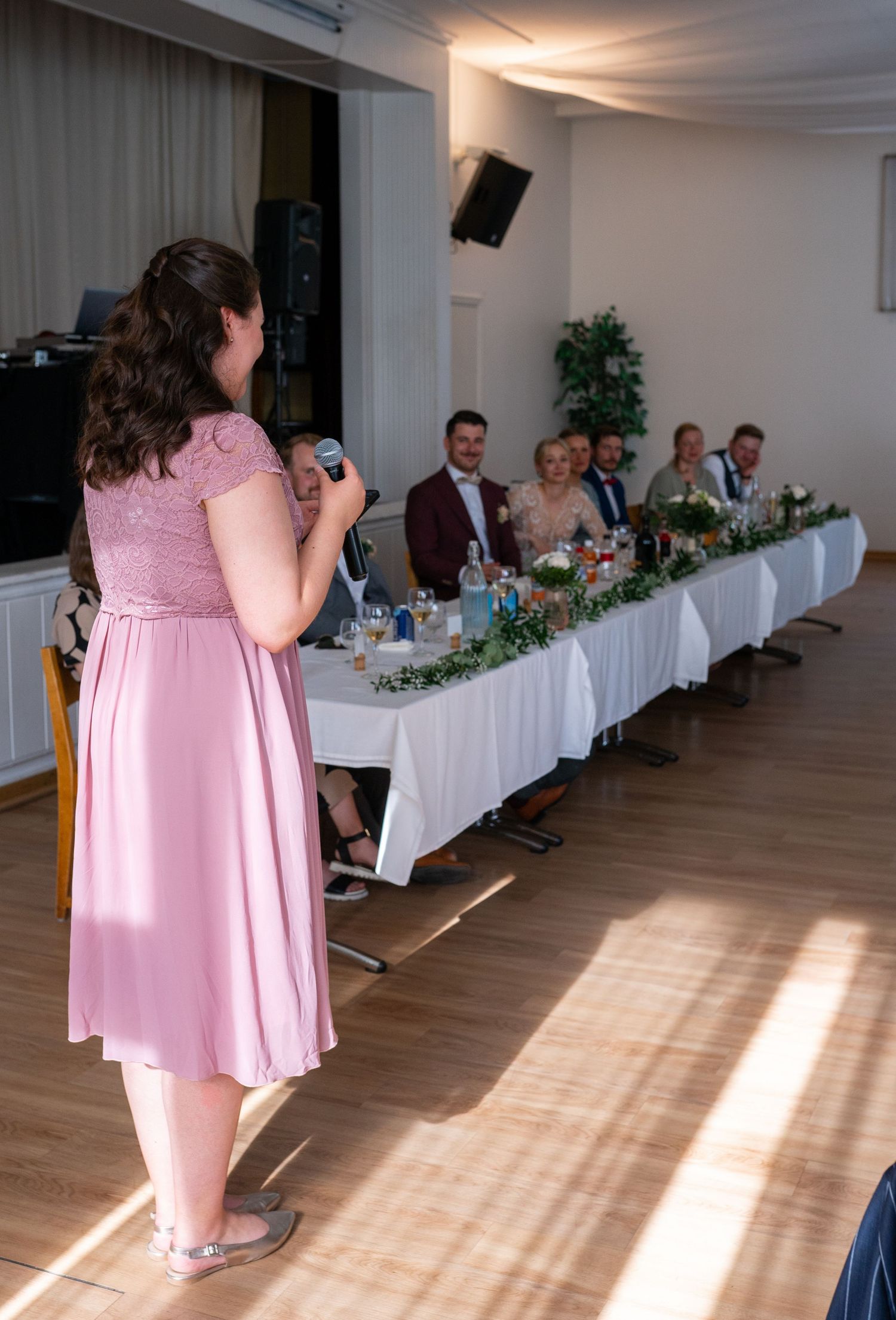 Woman in pink dress giving a speech at a wedding reception before the head table.