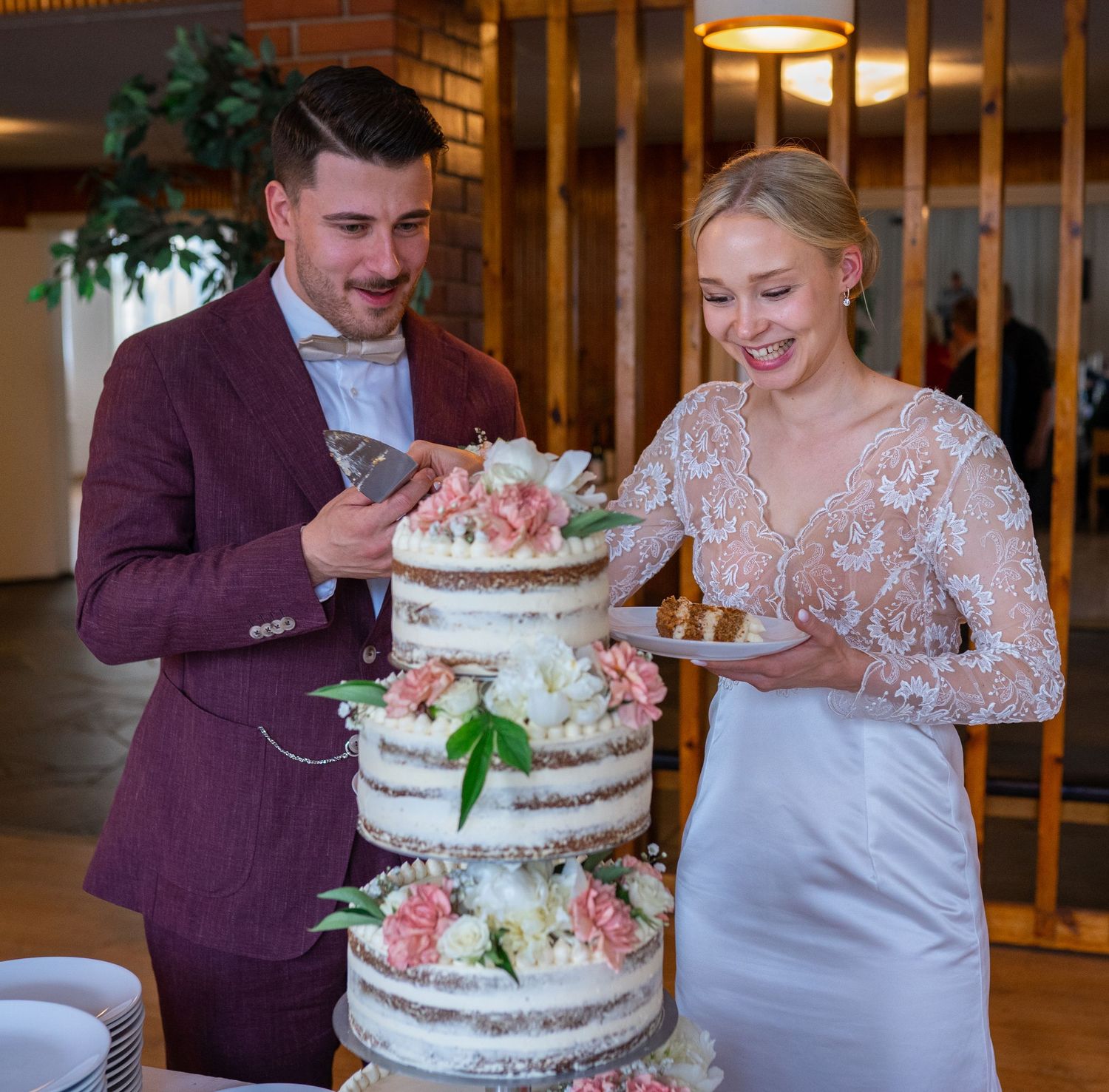 Smiling bride and groom in burgundy suit cutting their three-tier naked wedding cake together.