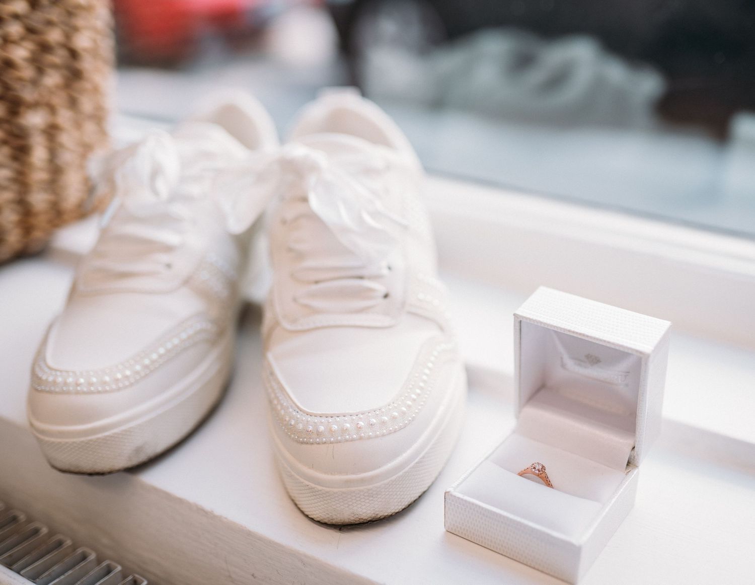 White bridal sneakers beside an open ring box with a gold engagement ring on a windowsill.