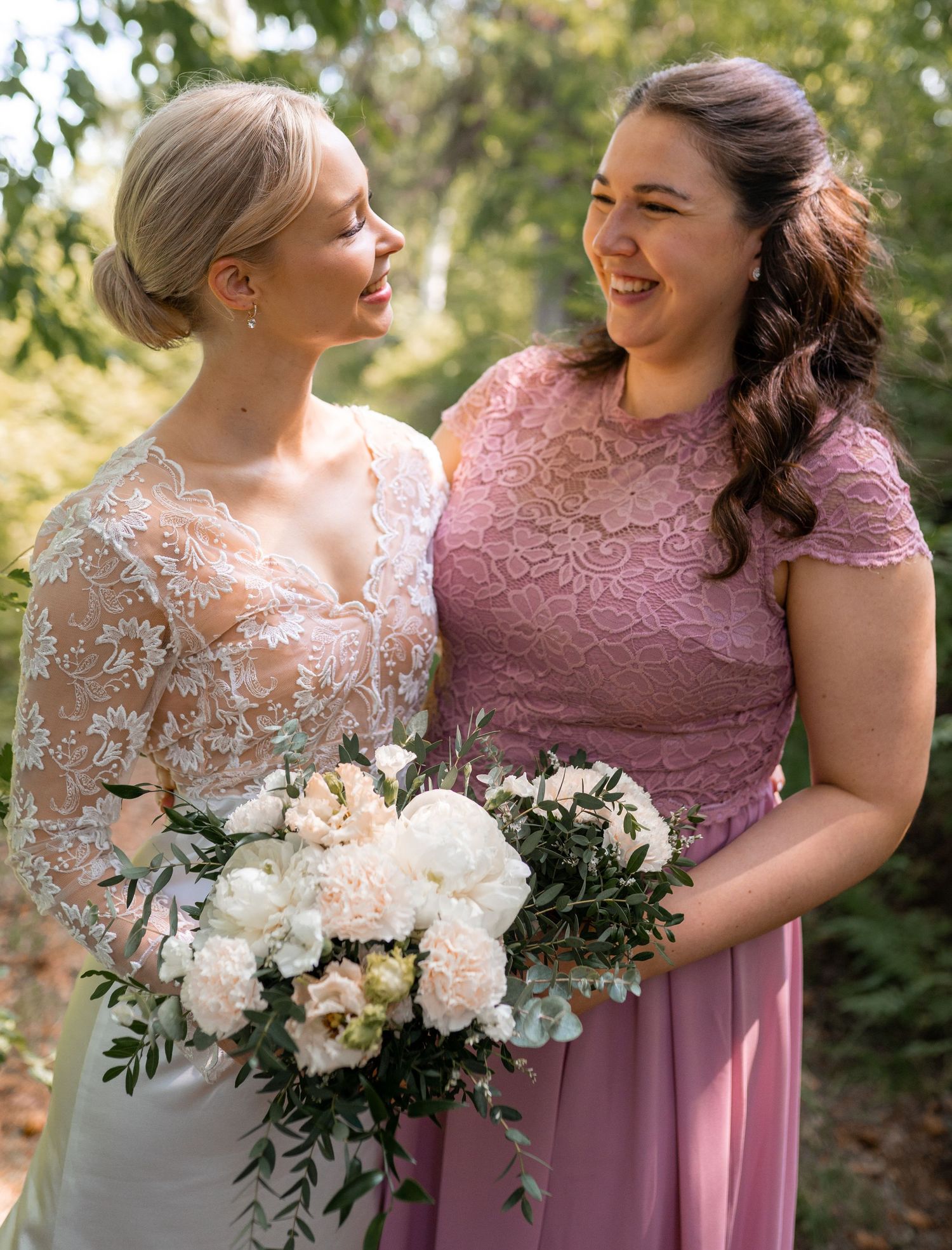 Bride in lace wedding gown and bridesmaid in pink lace dress holding white floral bouquets outdoors.