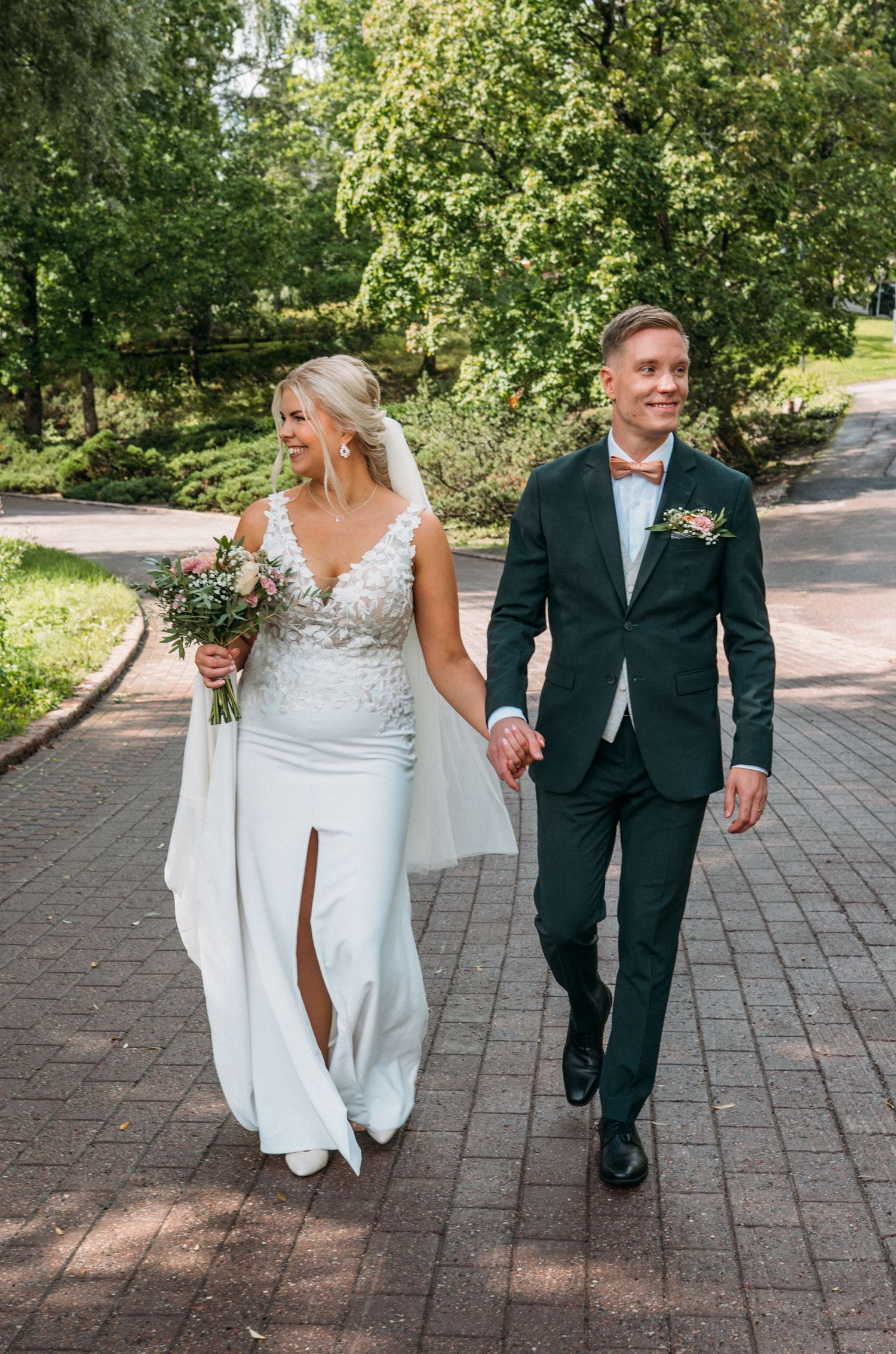 Elegant bride in a white lace gown and groom in a dark suit with bow tie walking through a sunny green park.