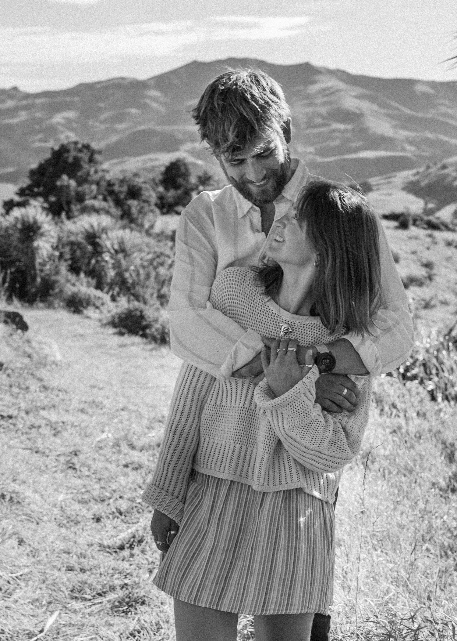 A couple laughs and hugs closely in a black and white portrait with mountain landscape behind them.
