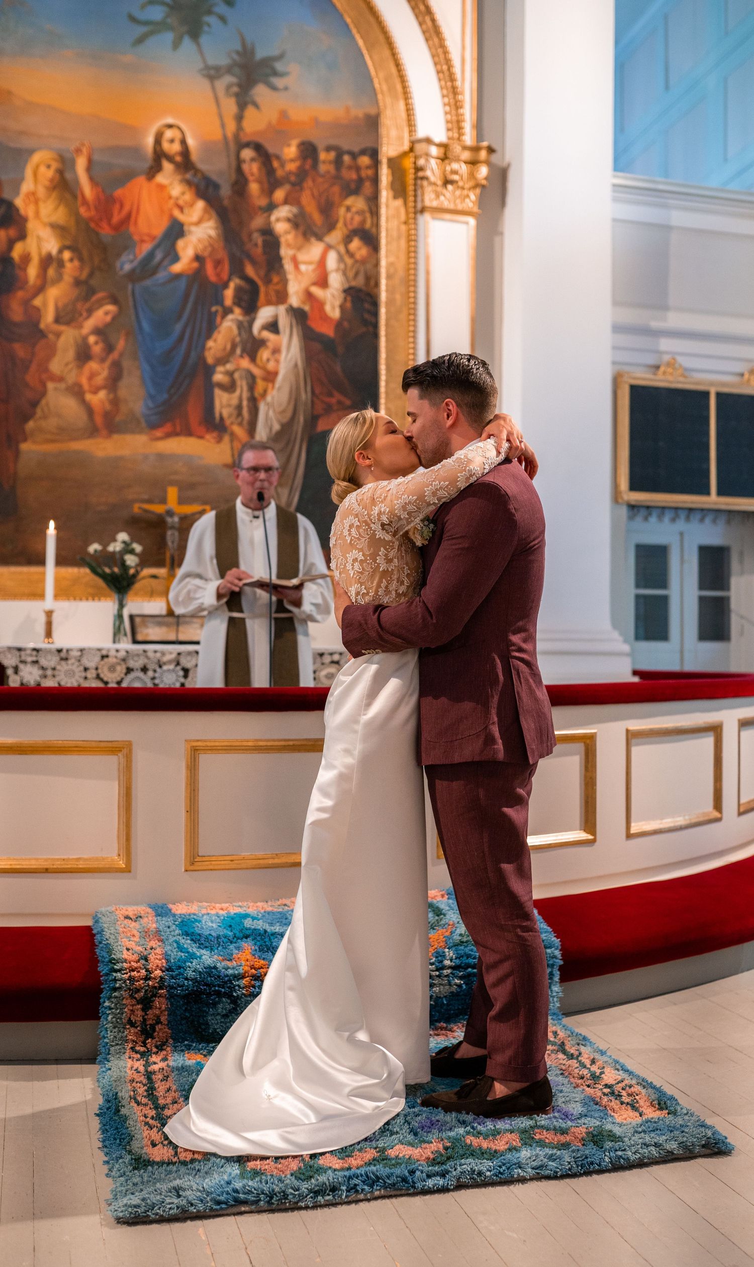 Bride and groom share first kiss at altar before large religious painting with officiant standing behind.