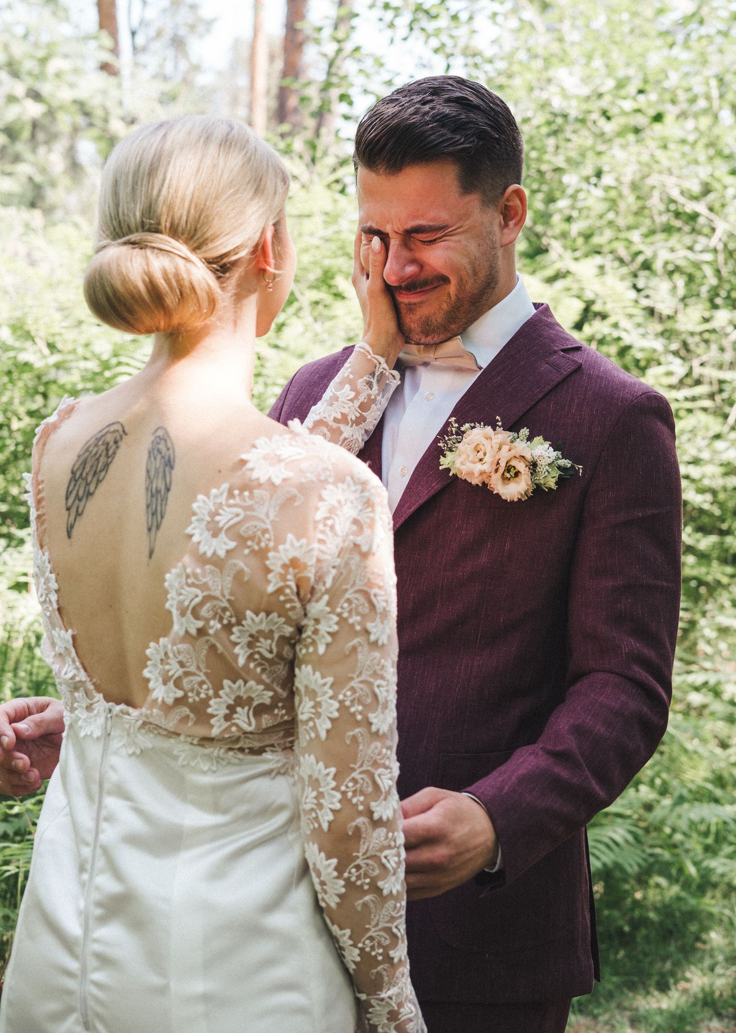 Emotional groom in burgundy suit wipes tears during first look with bride wearing lace gown in forest.