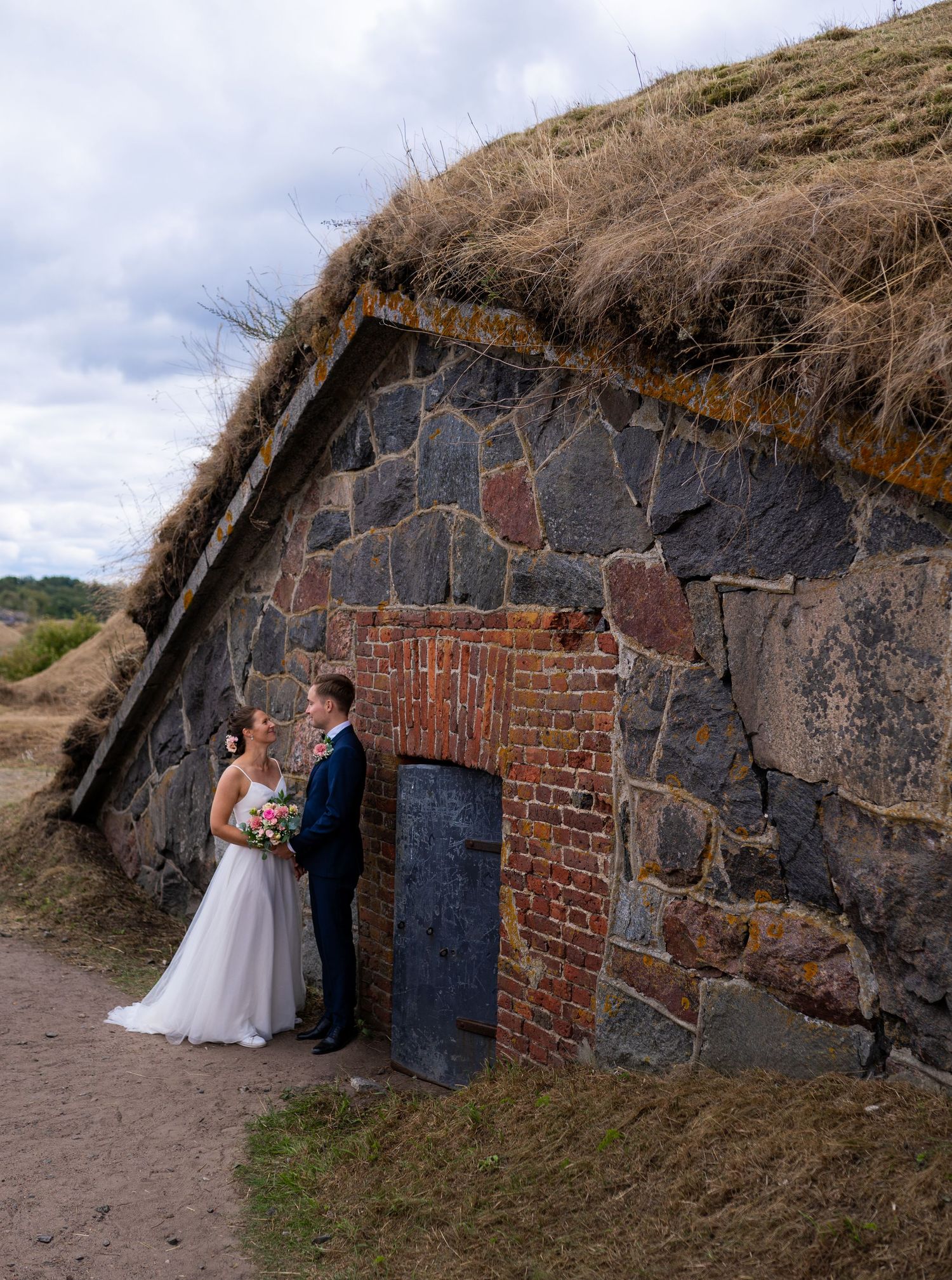 Bride and groom standing by a rustic stone and brick building with grass-covered roof on their wedding day.