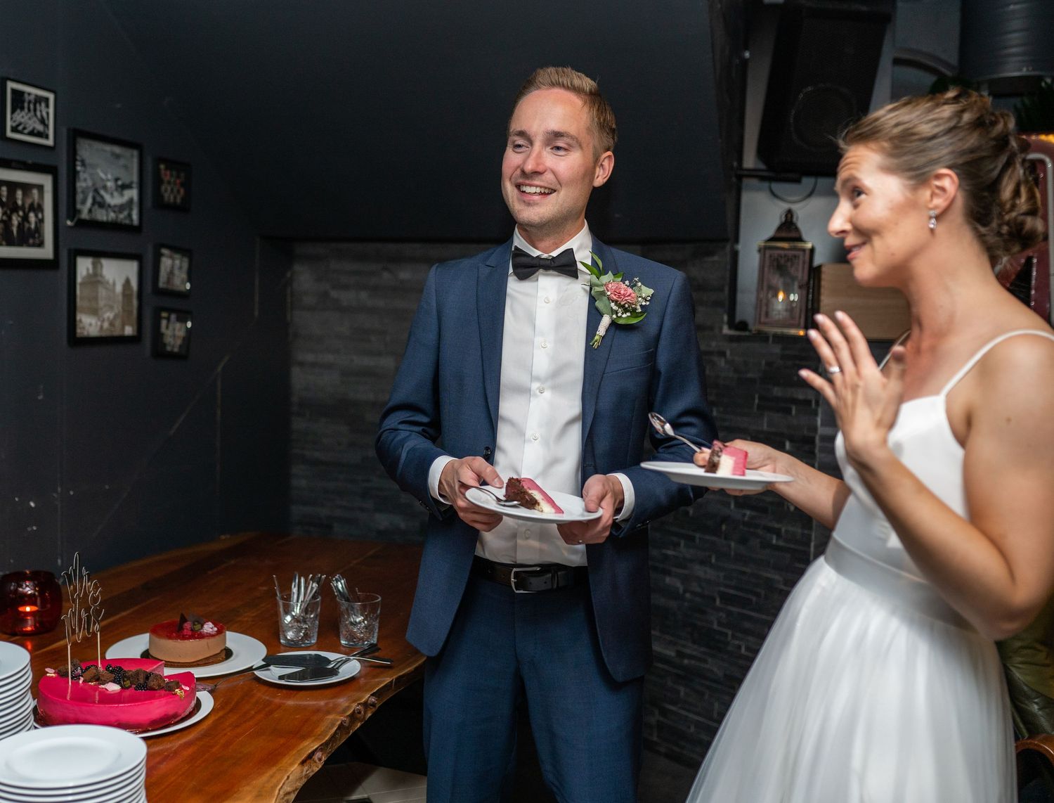 Happy wedding couple sharing cake slices, groom holding plate while bride claps joyfully.