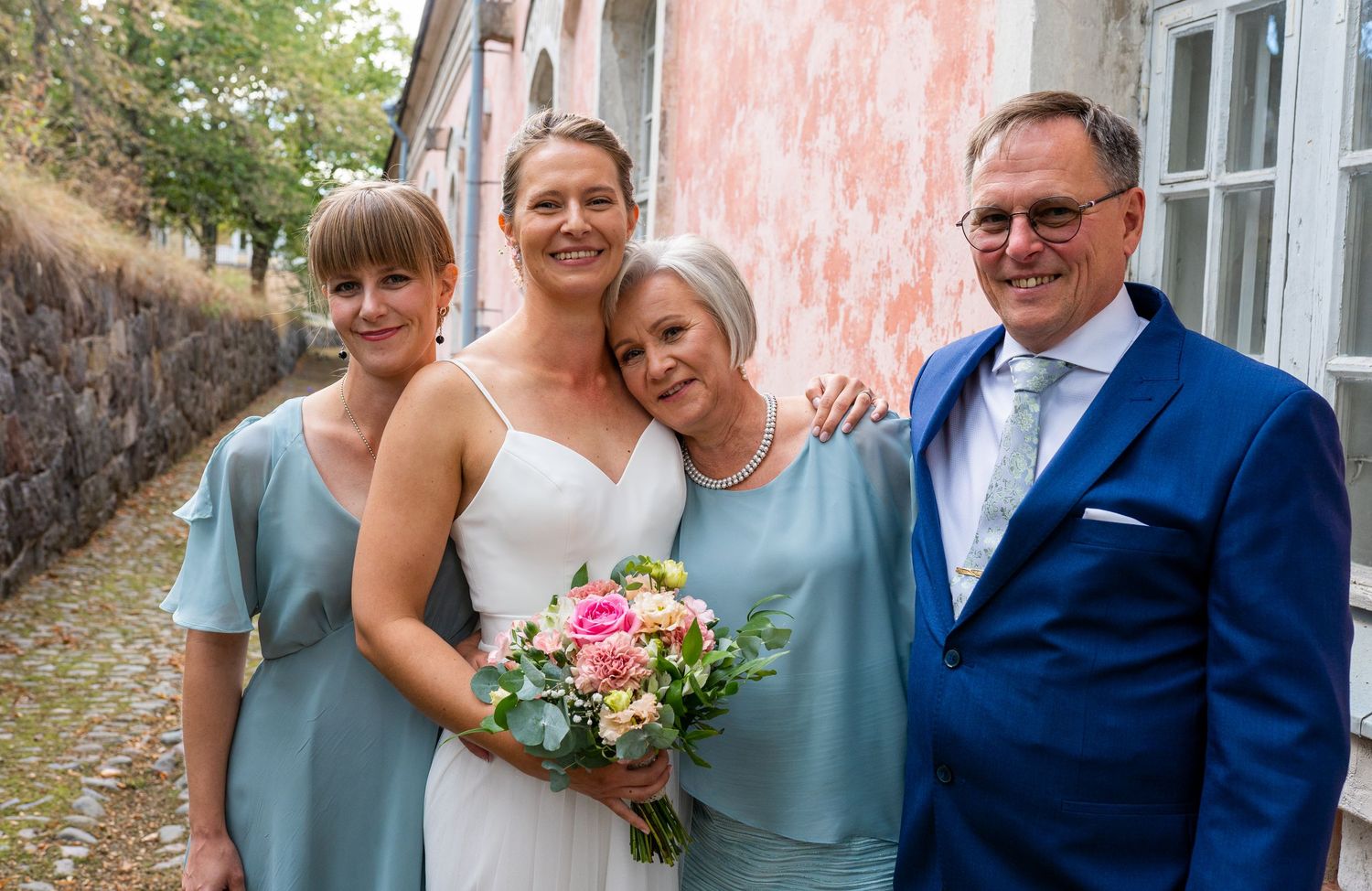 Bride in white gown with pink bouquet smiles with three family members in teal and blue attire outdoors.