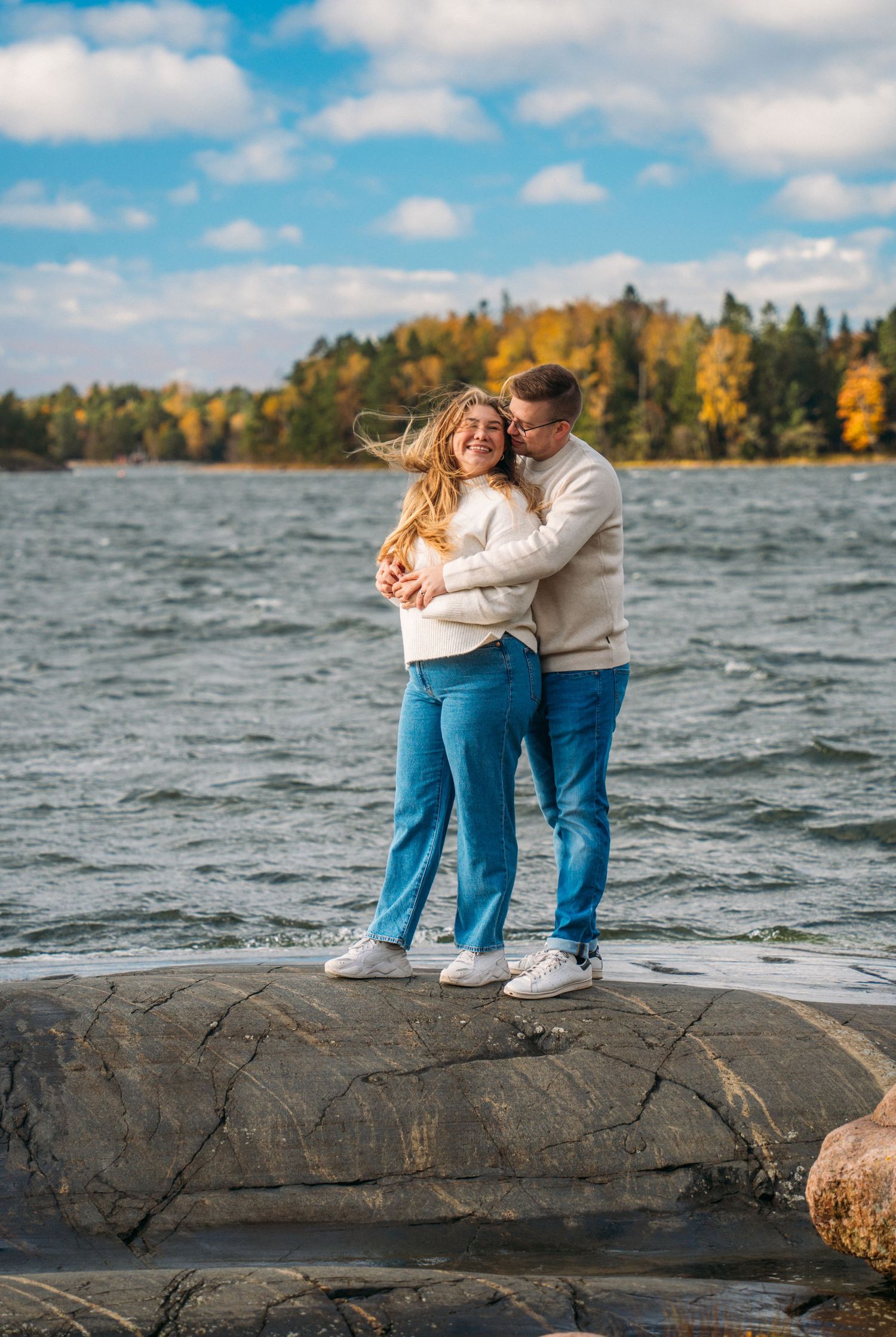 Couple embracing on rocky shoreline with autumn trees and blue sky in background.