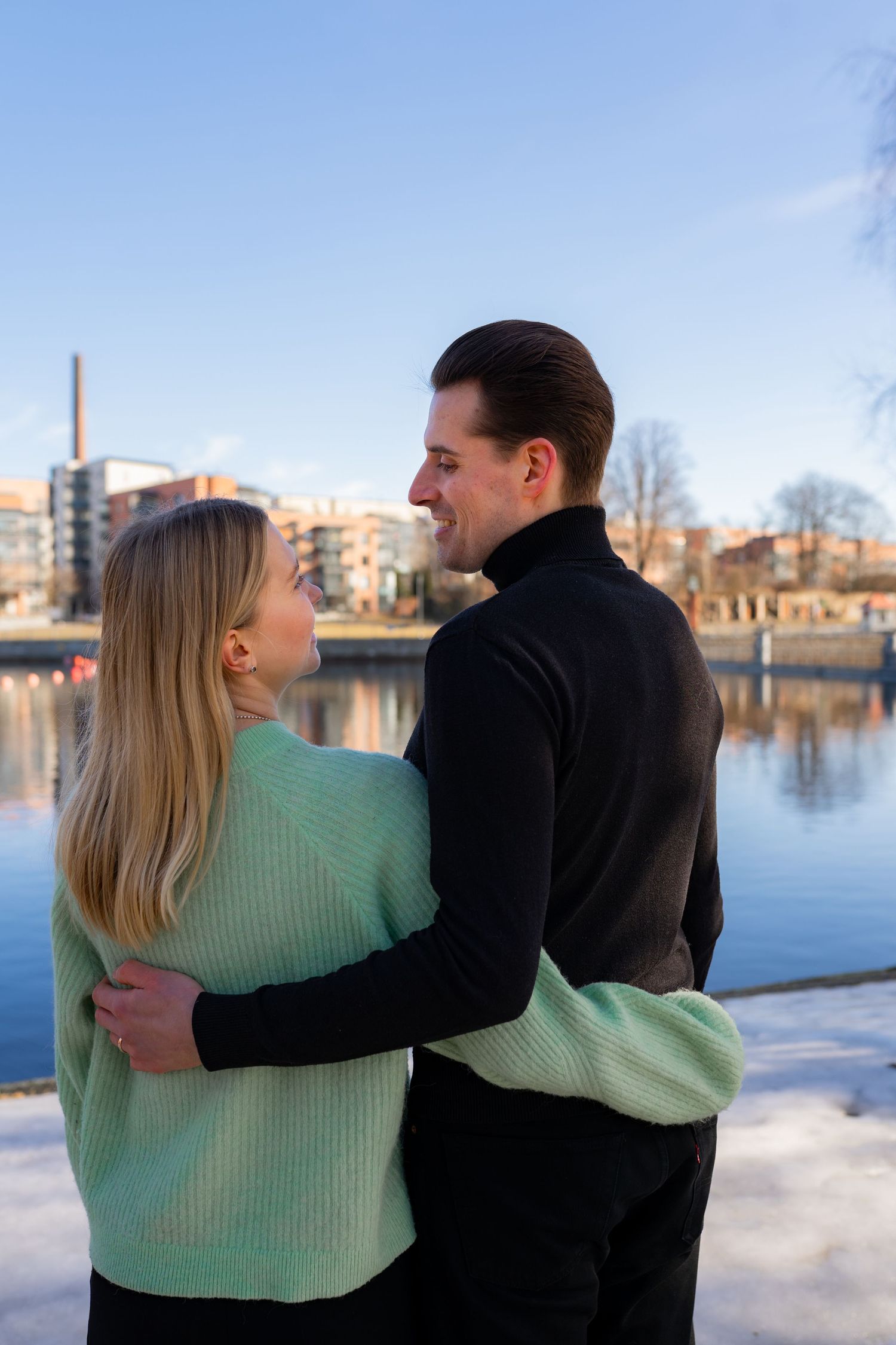 A smiling couple embraces by a riverside in winter, the woman in mint green and man in black turtleneck.