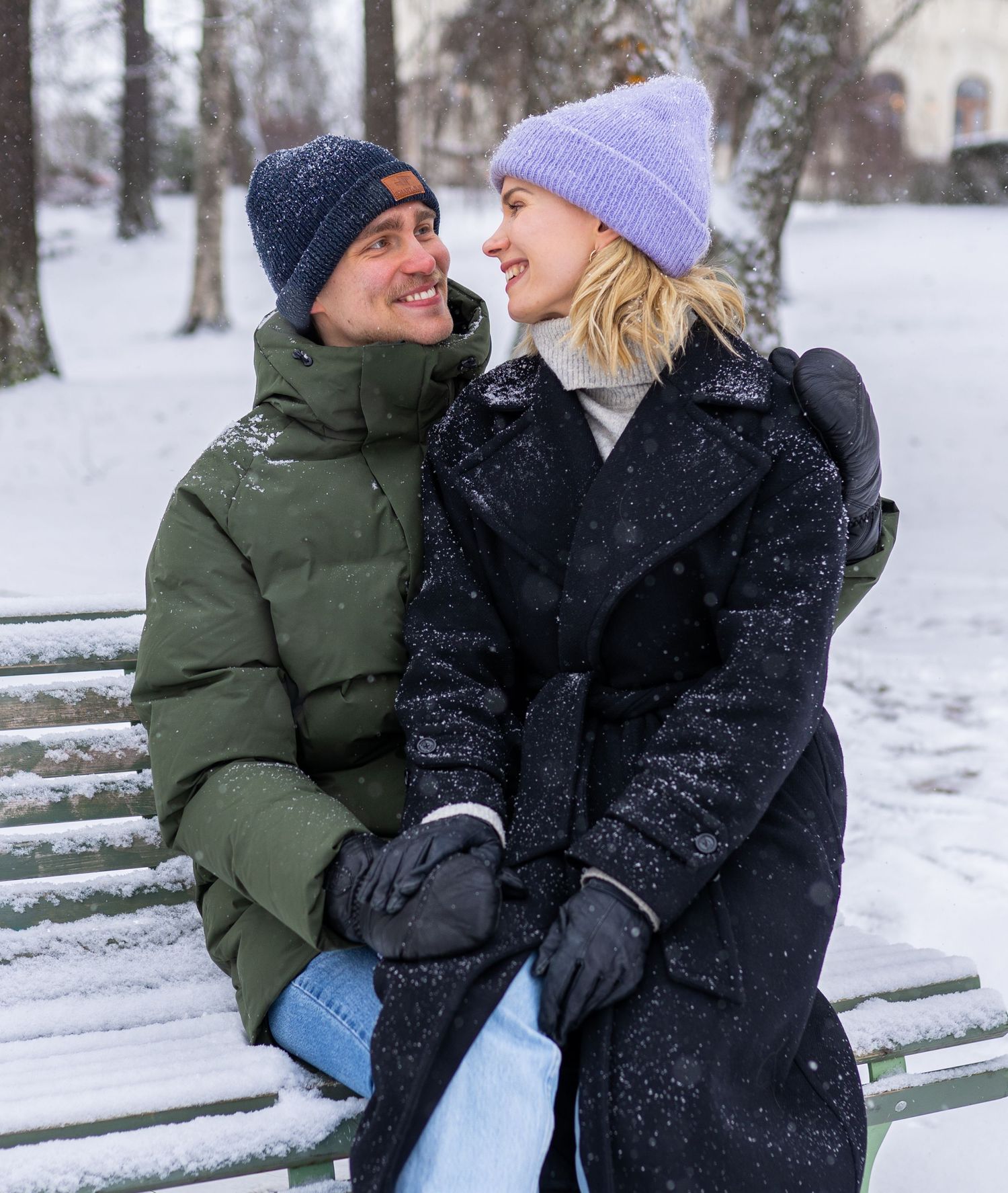 Couple sitting on snowy bench in winter park, wearing warm coats and knit hats, smiling at each other.
