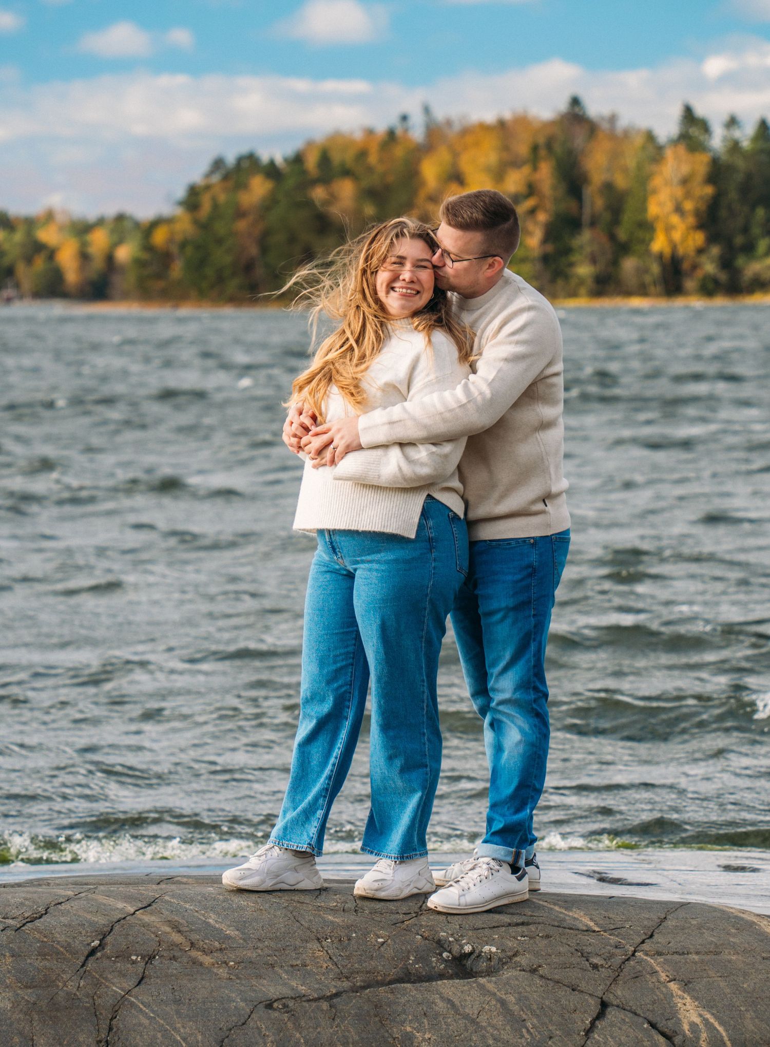 A couple embraces on rocky shore by a lake, wearing matching white sweaters and jeans, with autumn trees in background.