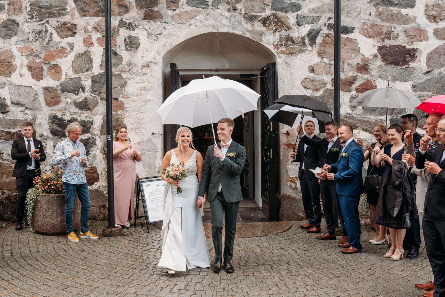 Newlyweds walk under white umbrella as guests applaud outside a rustic stone building on rainy day.