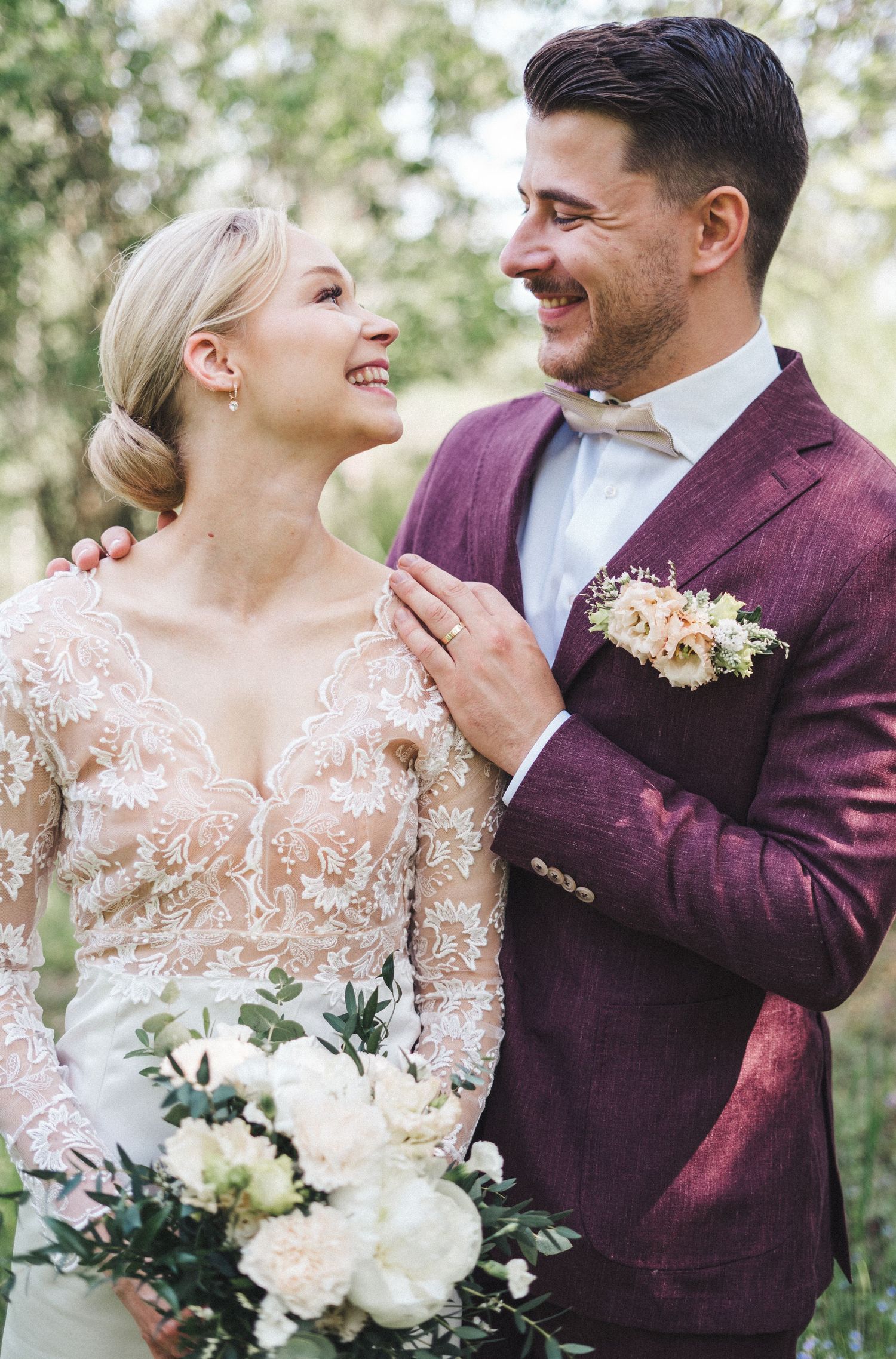Smiling bride and groom in burgundy suit share a joyful moment outdoors with bouquet.
