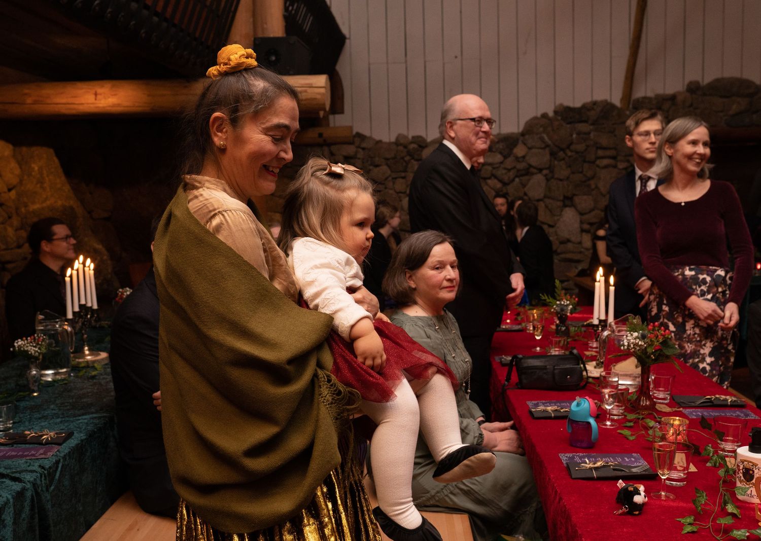 Family gathering at a candlelit medieval banquet, woman holding toddler in white dress near decorated table.