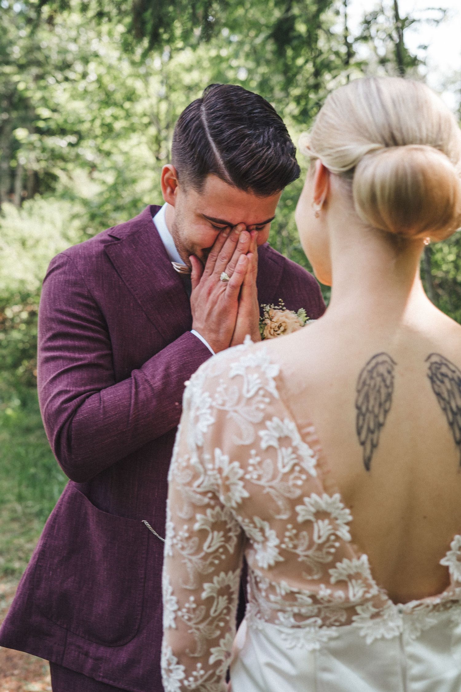 Emotional groom in burgundy suit covers face with hands while crying during first look with bride in lace wedding dress.