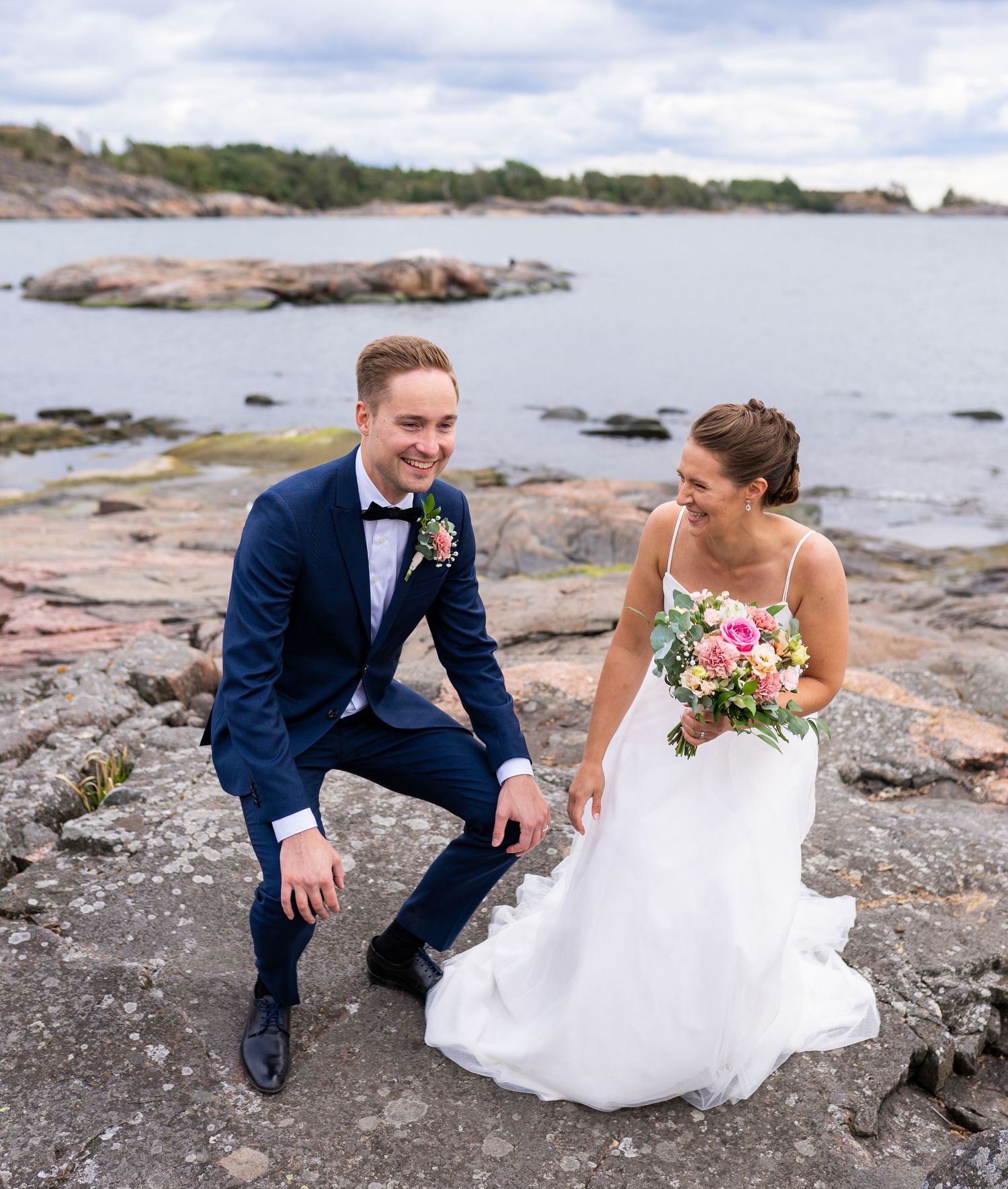 Laughing bride and groom sitting on coastal rocks, she holds pink bouquet, groom in navy suit with bow tie.