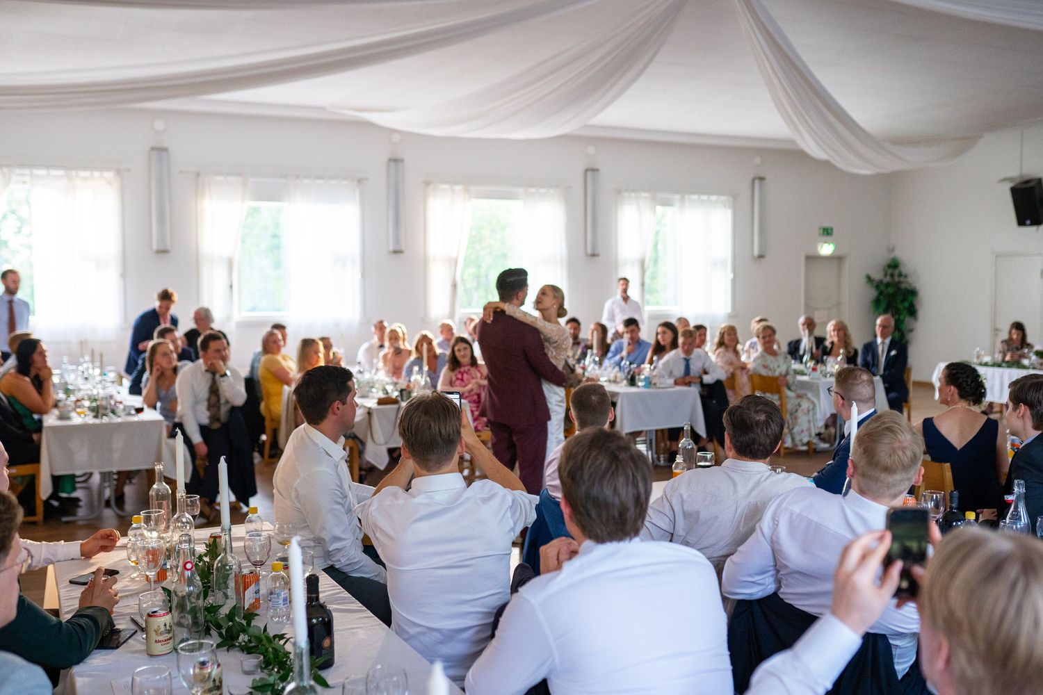 Wedding reception speech moment with guests seated at decorated tables in an elegant white draped venue.