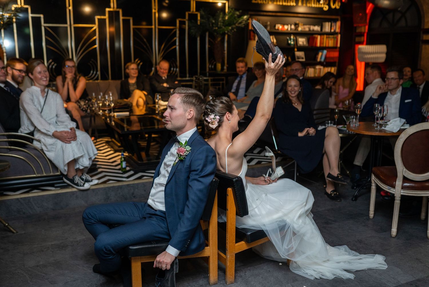 Bride holding up shoe during fun wedding shoe game while groom sits back to back.