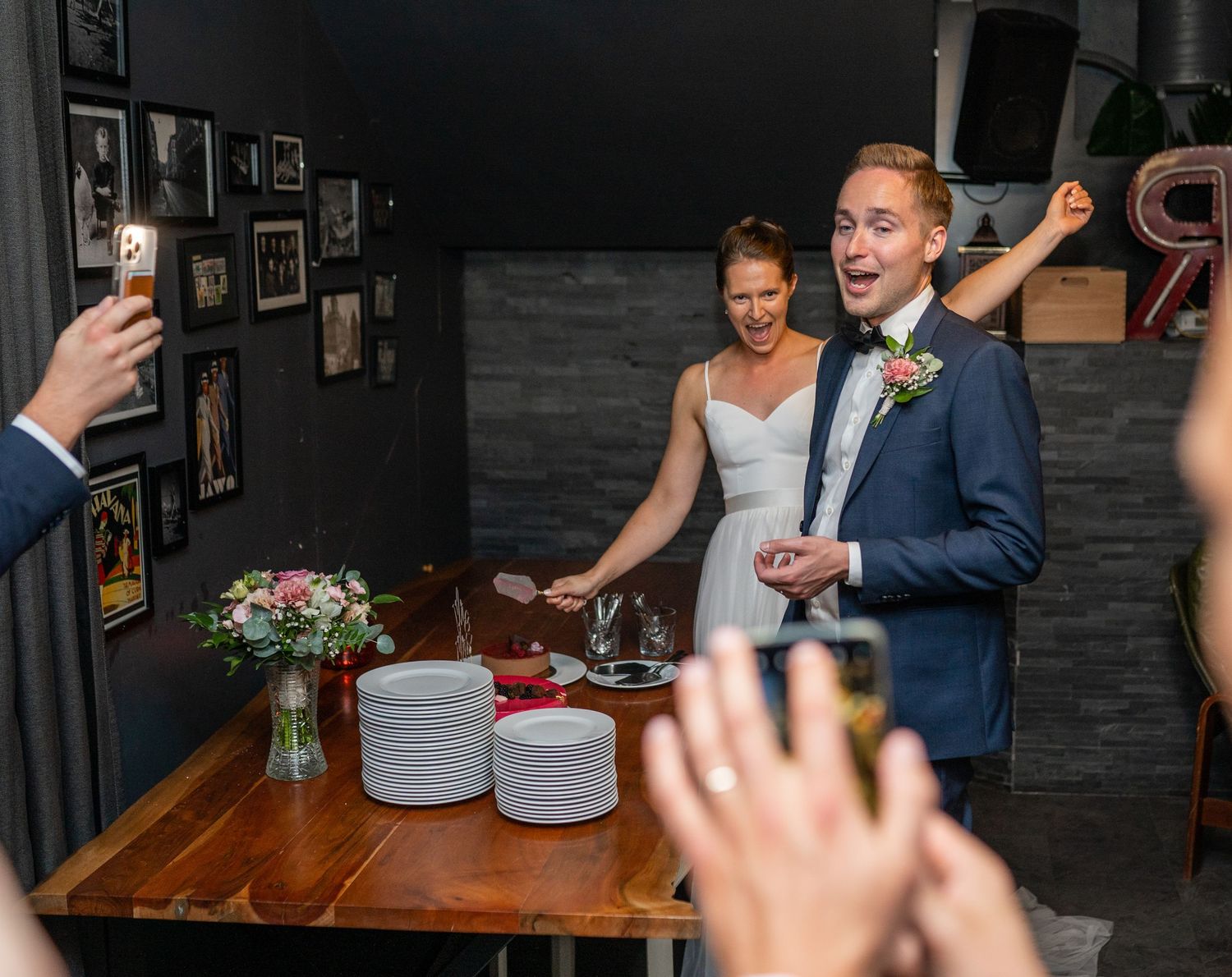Groom reacting joyfully beside bride at wedding cake table while guests take photos.