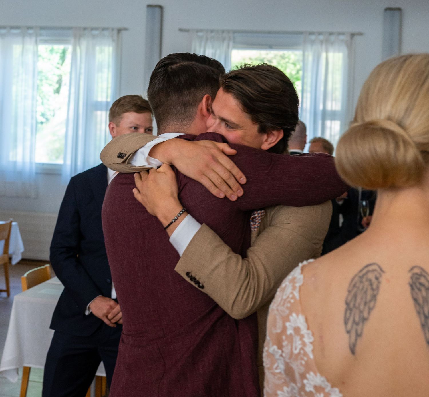 Two men share a warm hug at a wedding reception in a bright indoor venue.