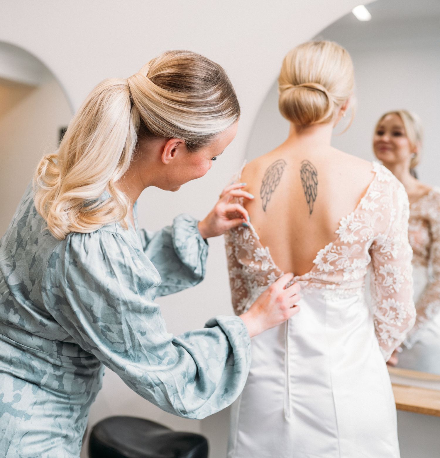 Blonde bridesmaid fastening buttons on the back of a brides lace wedding gown near a mirror.
