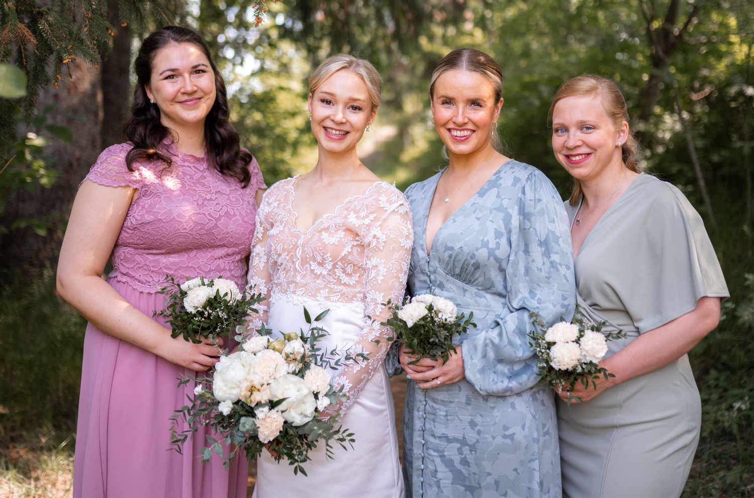 Bride in lace gown with three bridesmaids in pink, blue, and sage dresses outdoors.