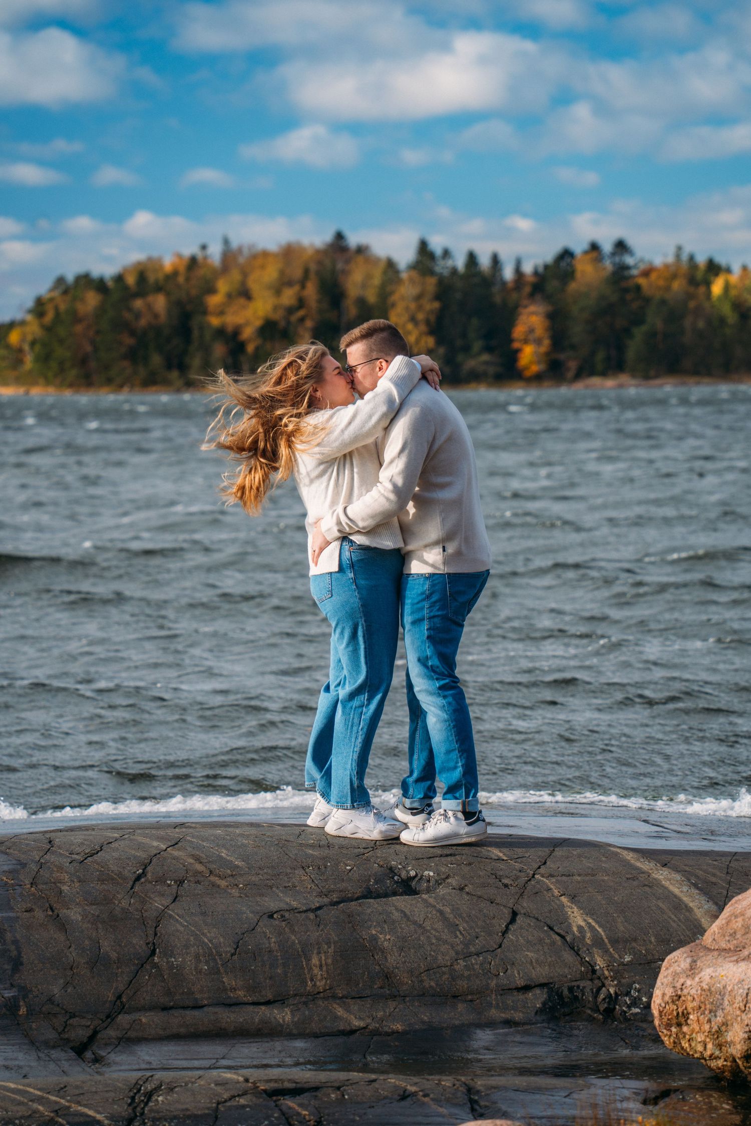 Couple kissing on rocky shoreline with autumn trees and blue sky in background.