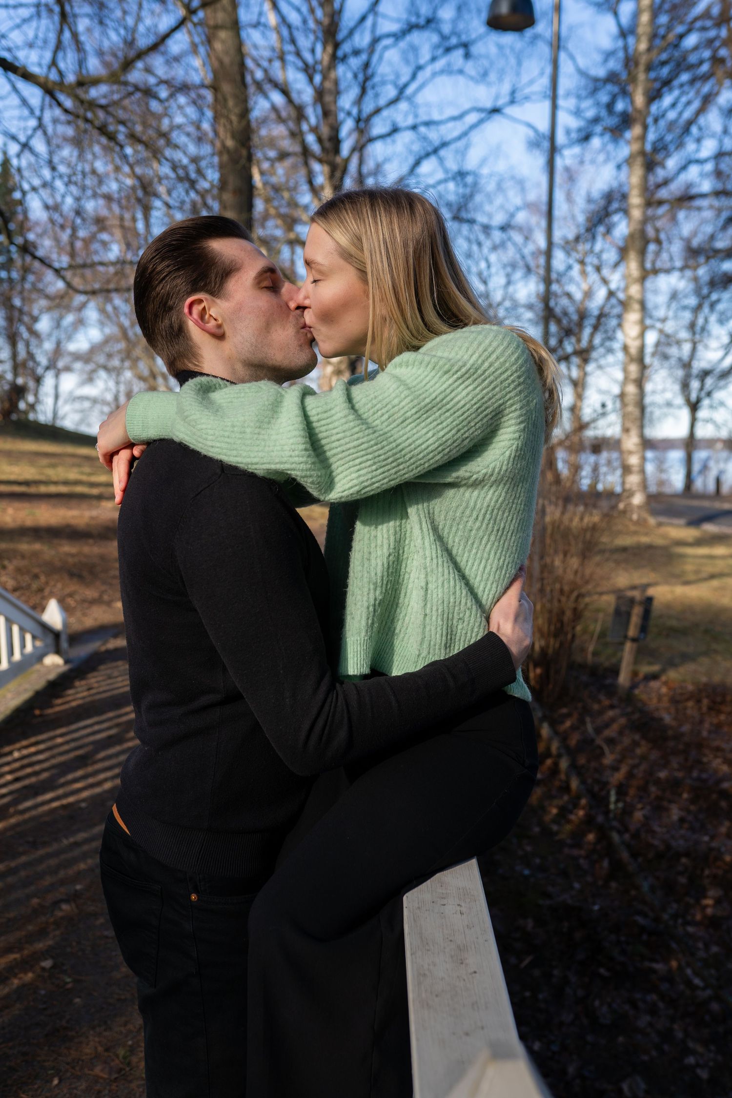 A couple sharing a romantic kiss outdoors in a park during autumn, man in black and woman in green sweater.
