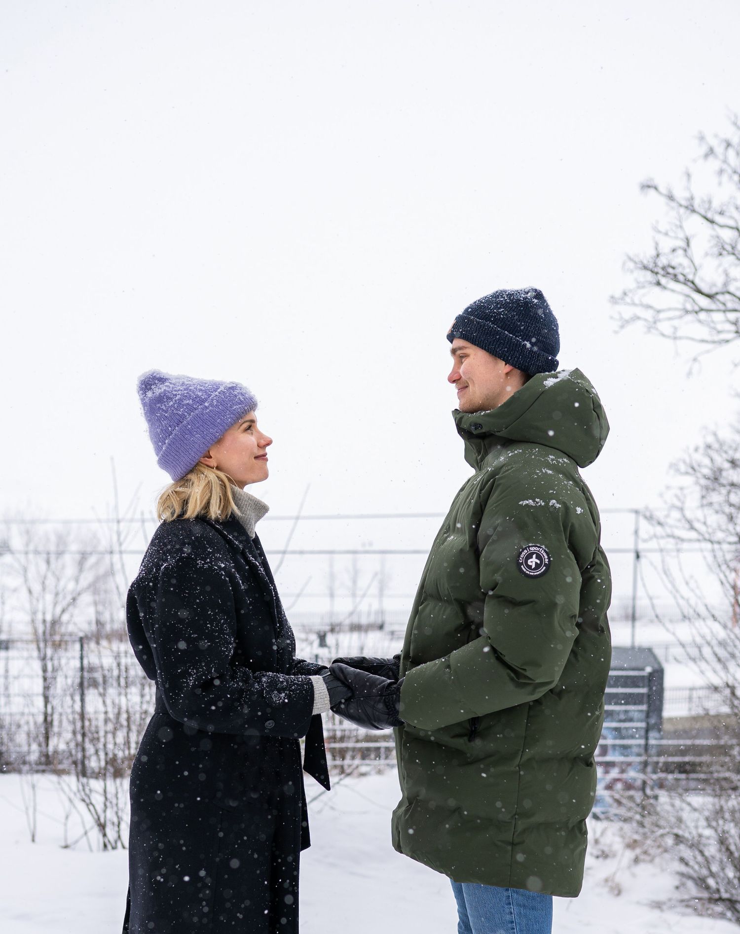 Couple holding hands in snowy winter landscape, wearing warm coats and knit hats while snowflakes fall around them.