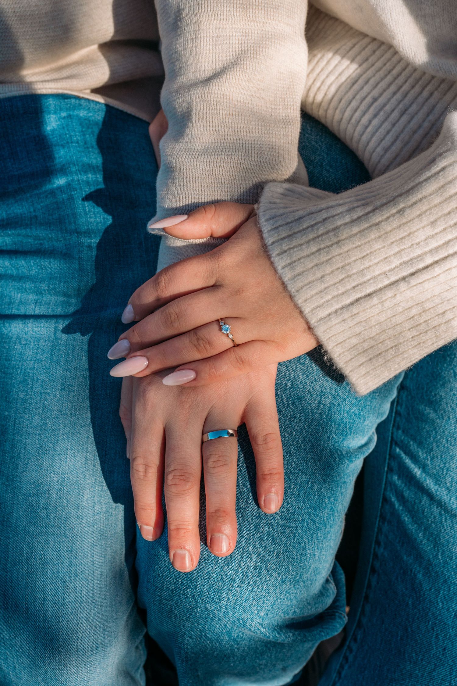 Close-up of two hands resting on blue jeans, one wearing a delicate ring, both in cream ribbed sweaters.