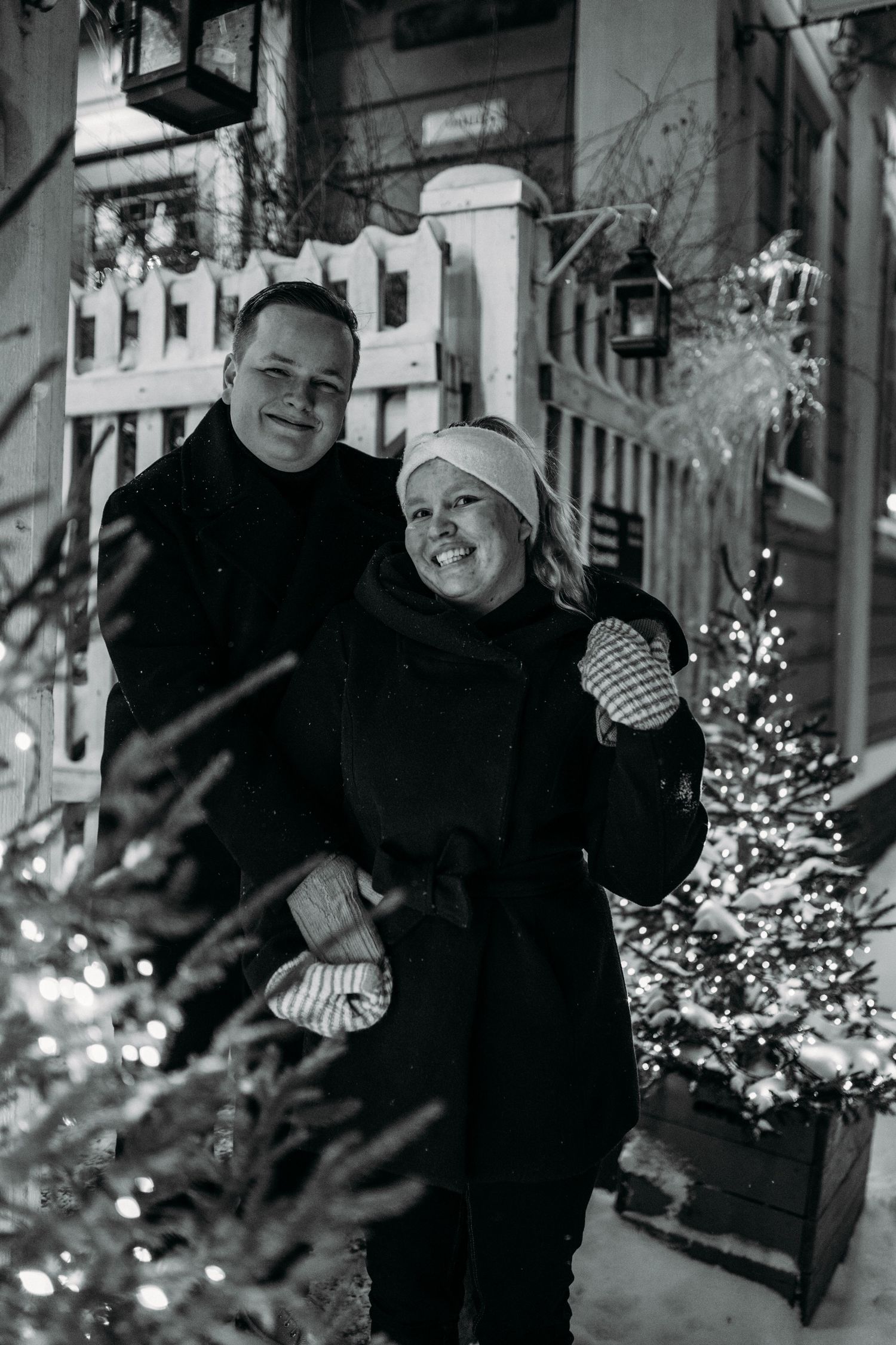 Black and white photo of a smiling couple in winter coats near Christmas trees with lights on a snowy evening.