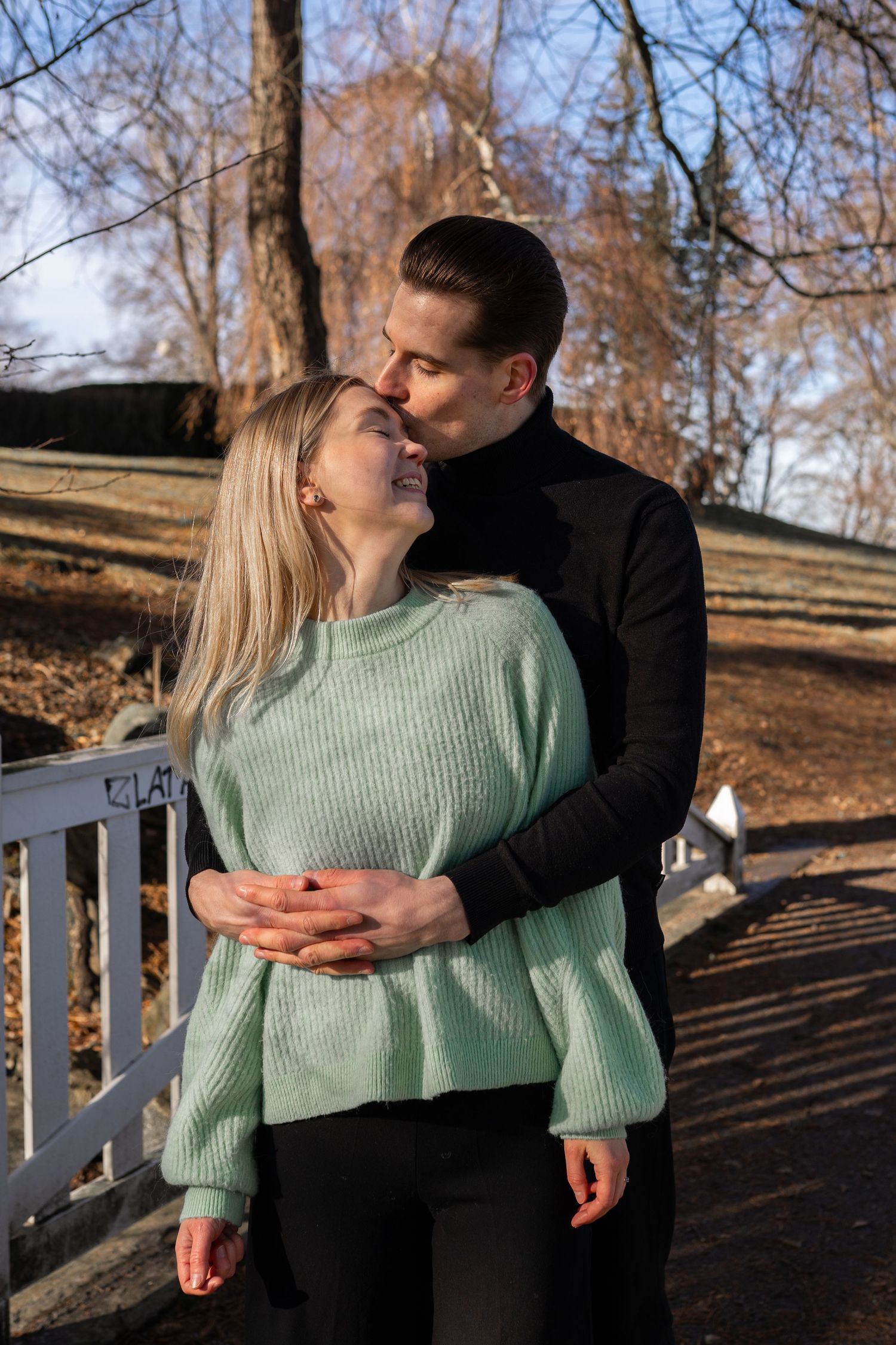 A couple embracing on a bridge outdoors, woman in mint sweater smiling up at man in black turtleneck kissing her forehead.