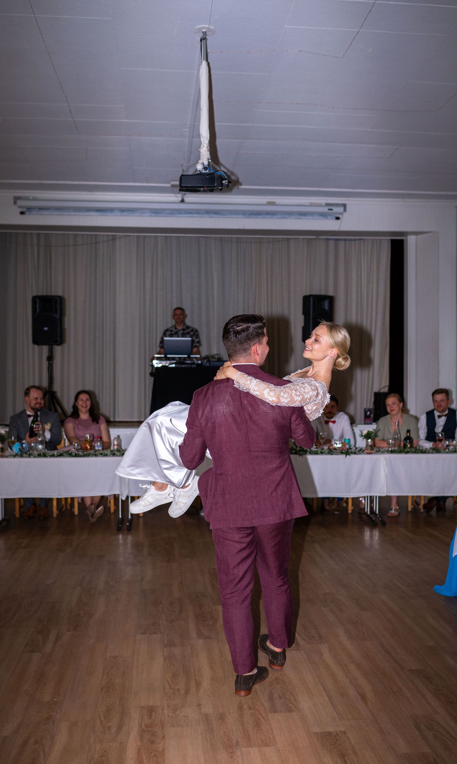 Groom lifts bride during first dance at wedding reception with DJ and guests visible in background.