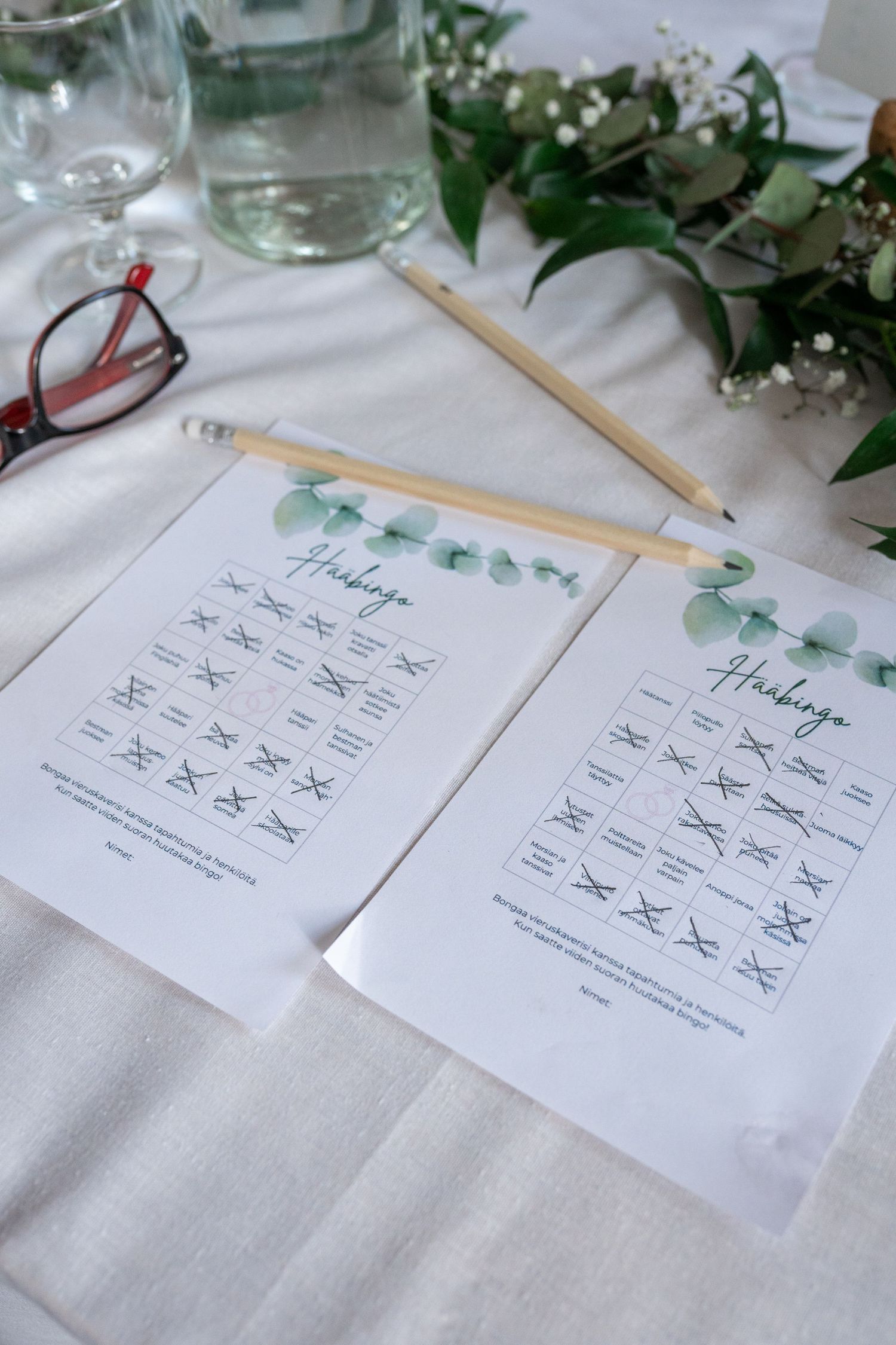 Wedding bingo game cards with eucalyptus design and pencil on white tablecloth.
