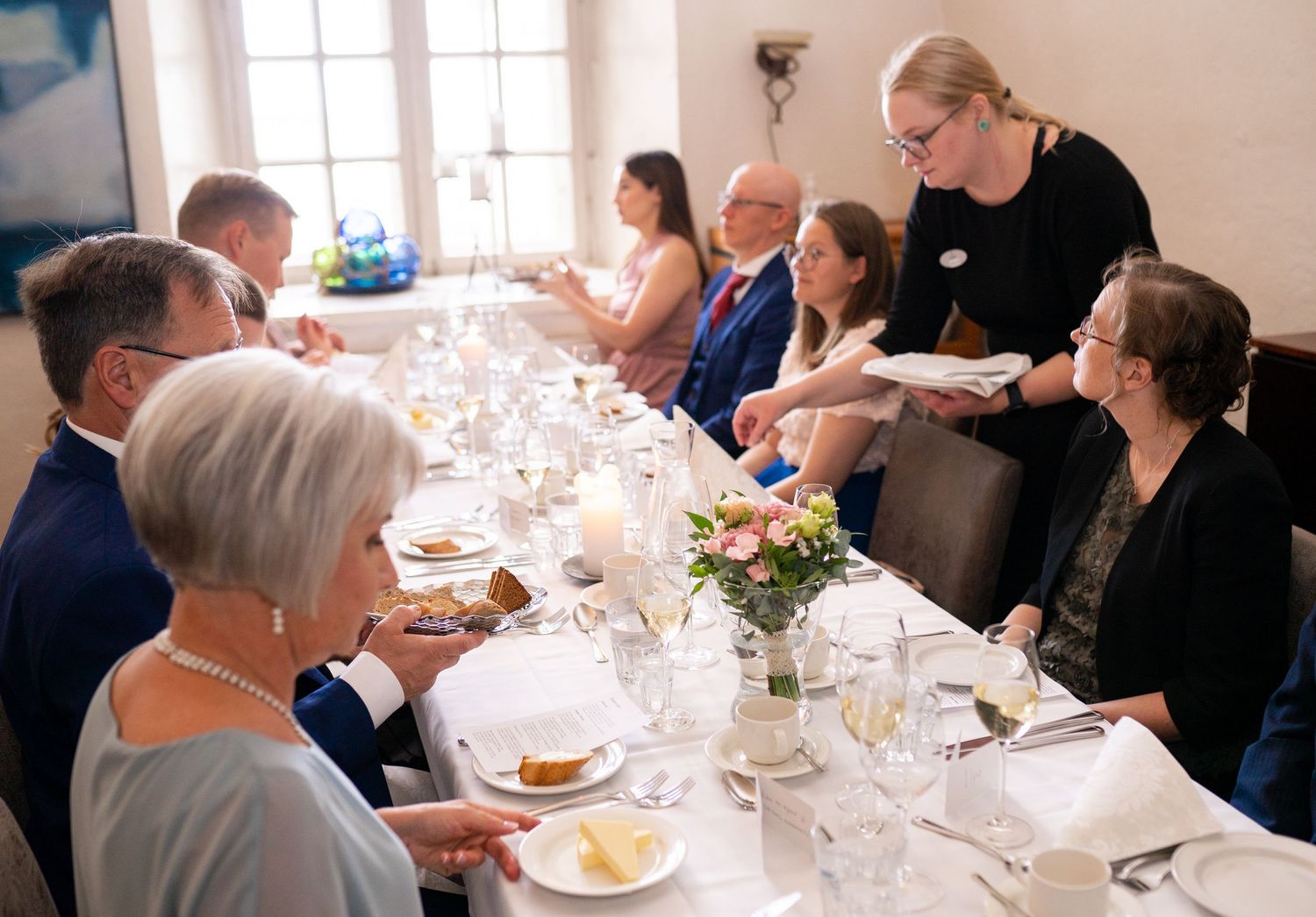 Server presenting dishes to wedding guests seated at a long formal dinner table with floral decor.