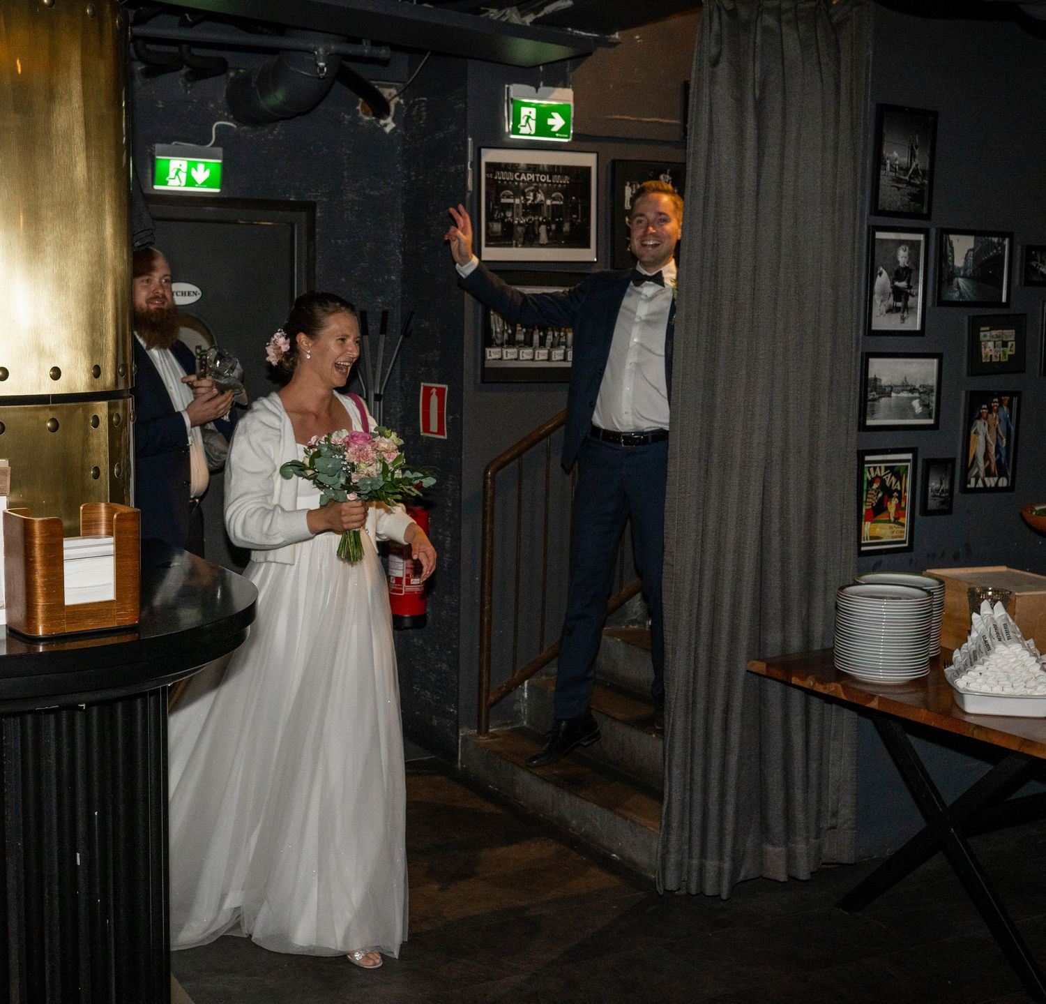 Bride in white gown holding bouquet enters venue as groom waves from staircase at wedding reception.