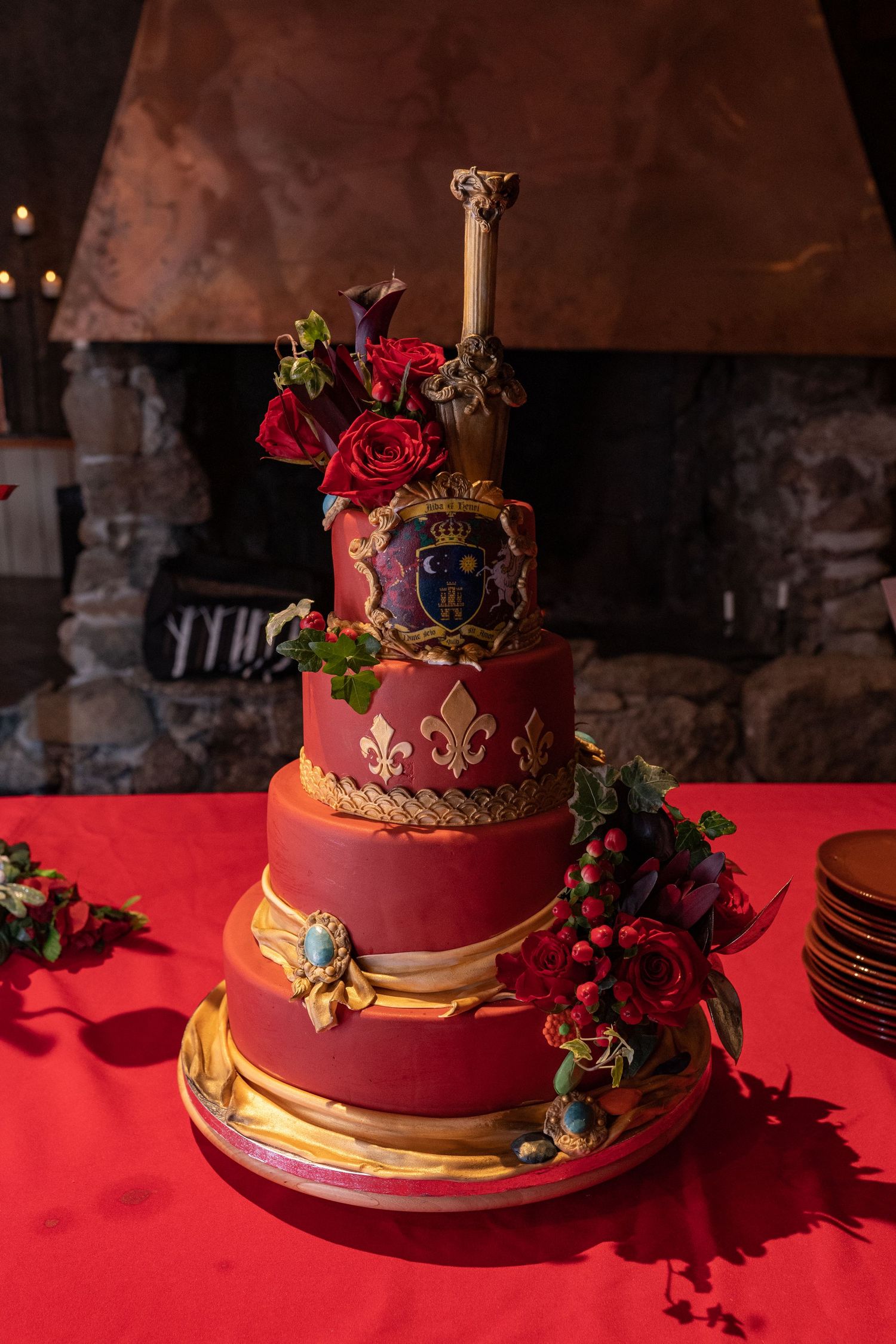 Elegant three-tier red and gold wedding cake with fleur-de-lis, roses, and a decorative sword topper by a stone fireplace.