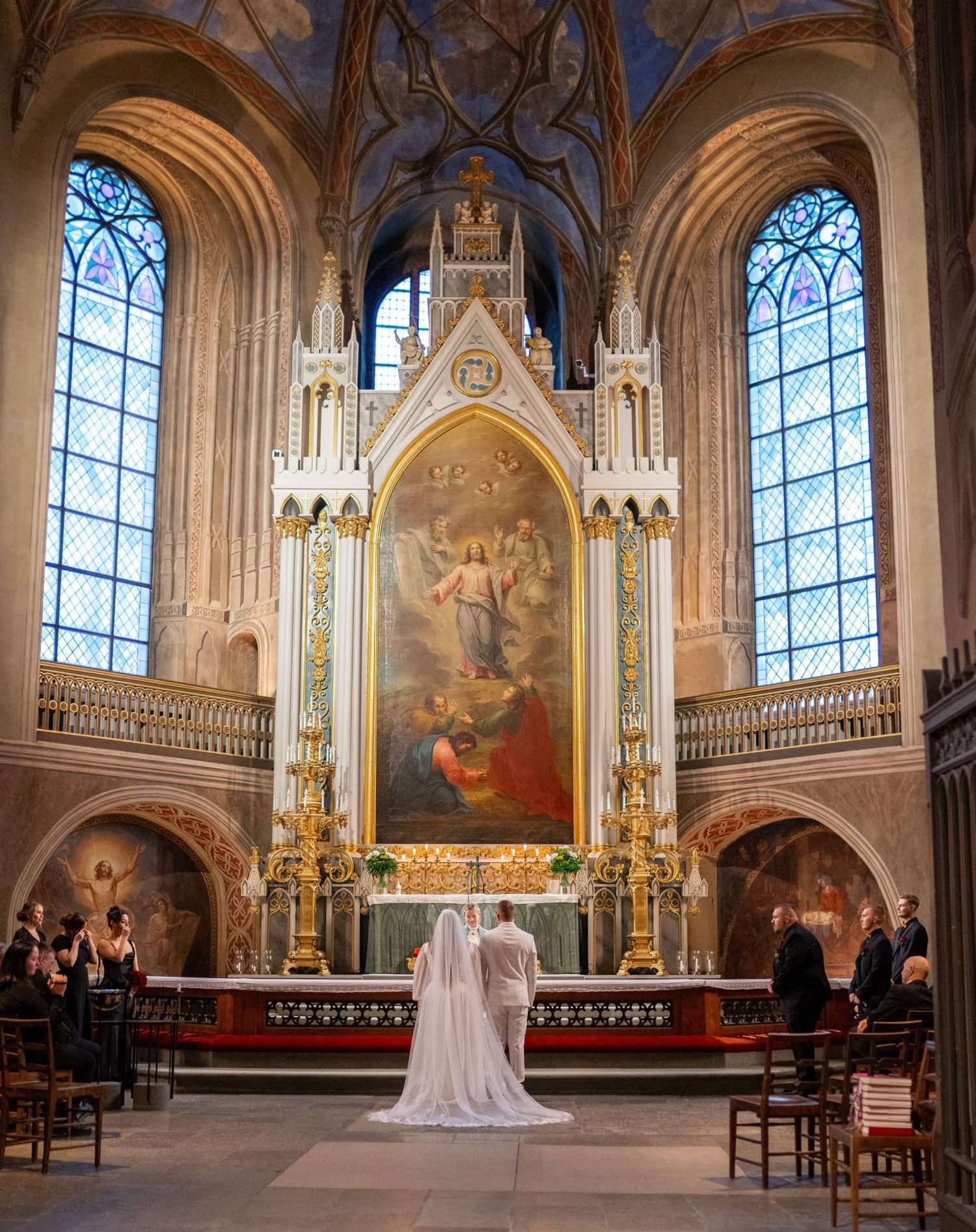 Bride in white veil standing before ornate golden altar in grand cathedral with stained glass windows.