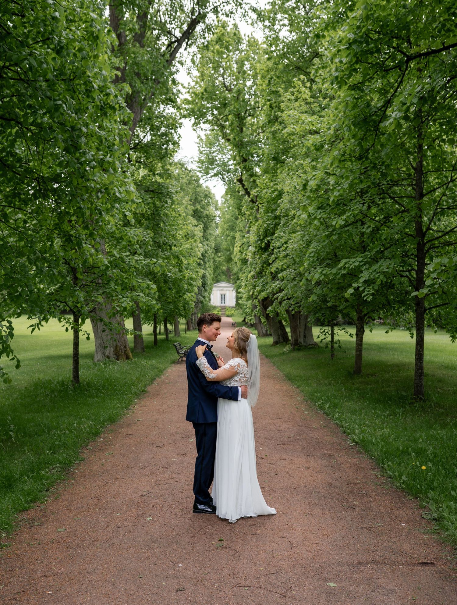 Bride and groom sharing a kiss on a tree-lined pathway in a lush green park on their wedding day.