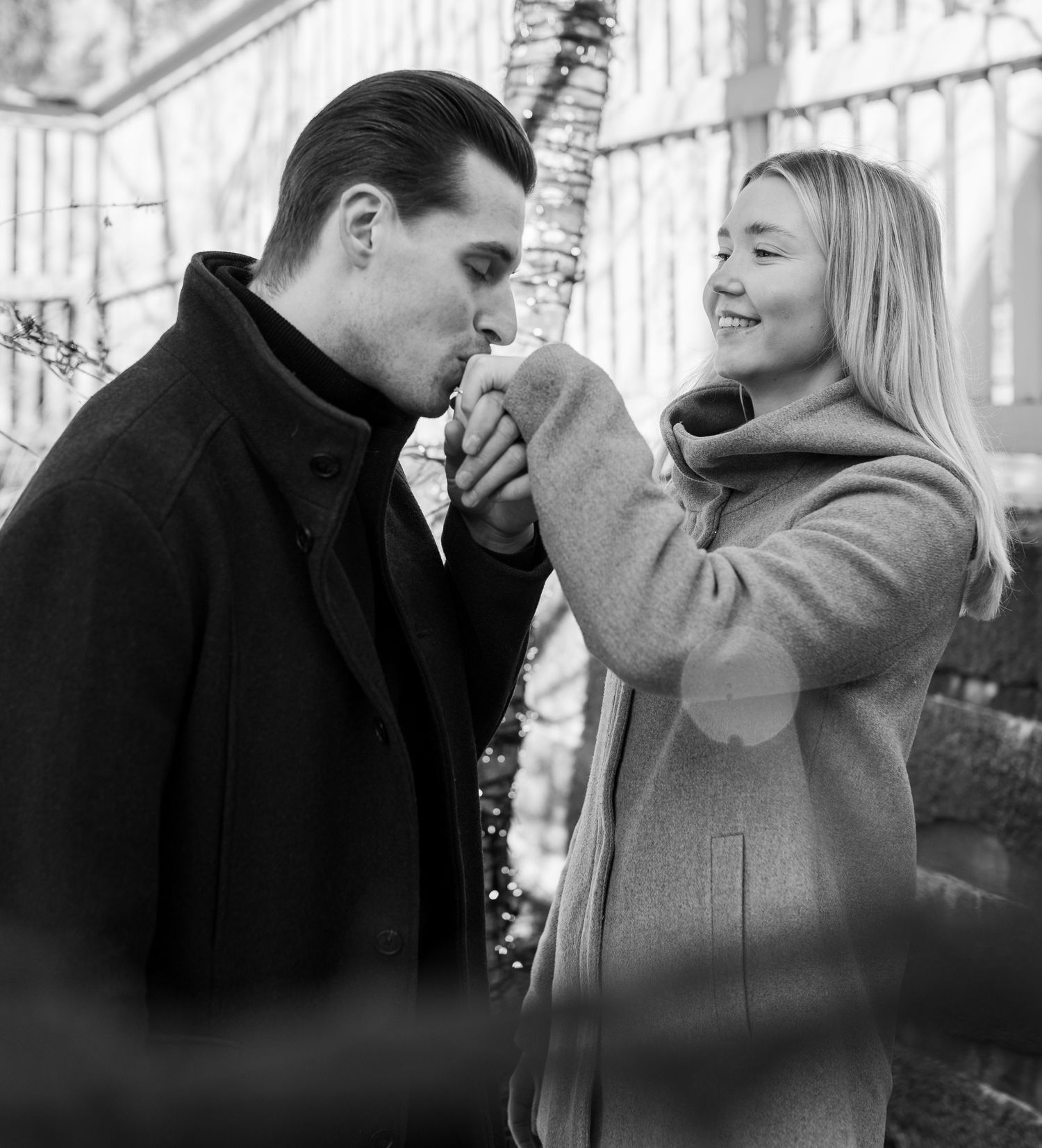 Black and white photo of a man kissing a smiling woman's hand outdoors in winter.