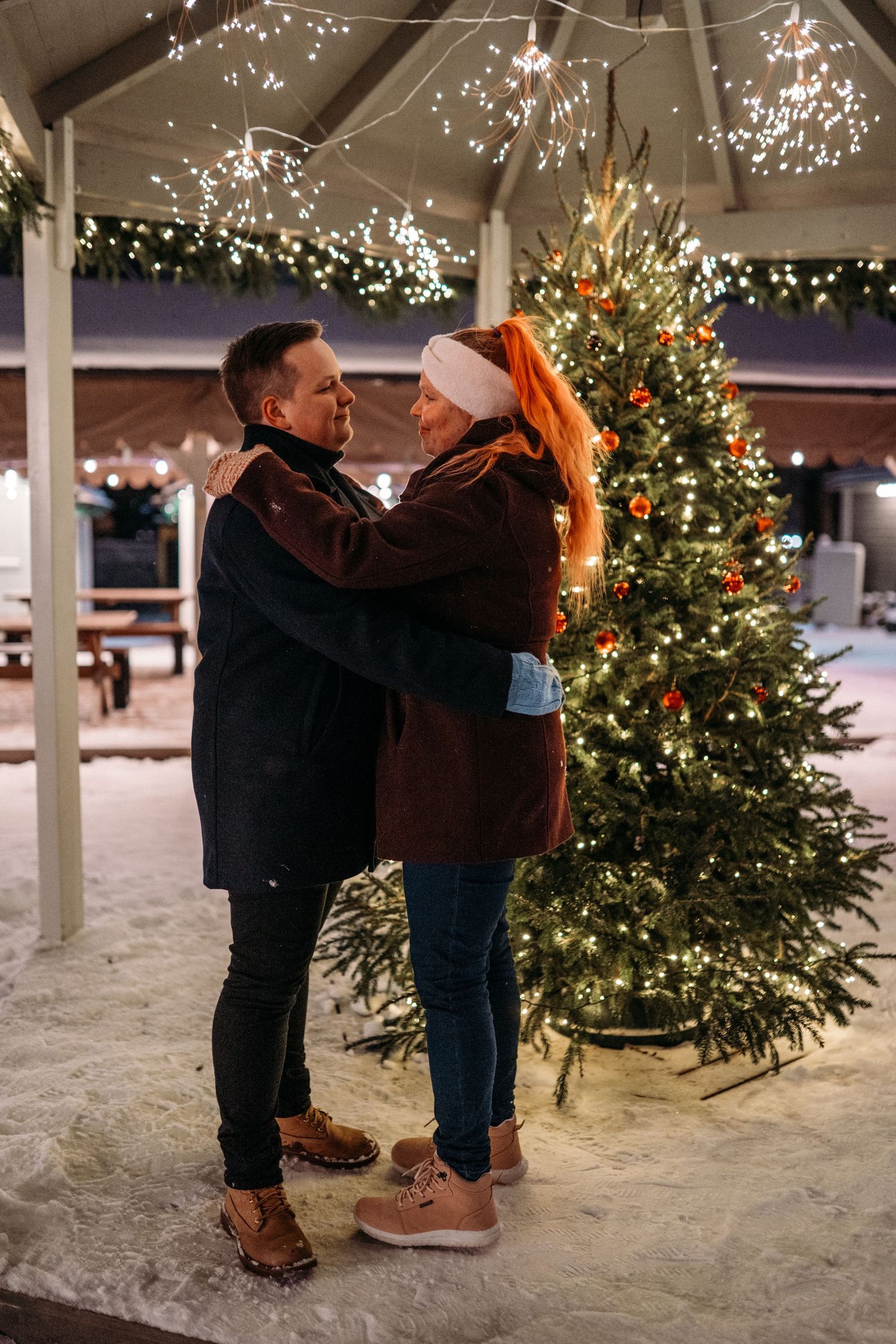 Couple embracing romantically in front of a decorated Christmas tree outdoors at night with festive lights.