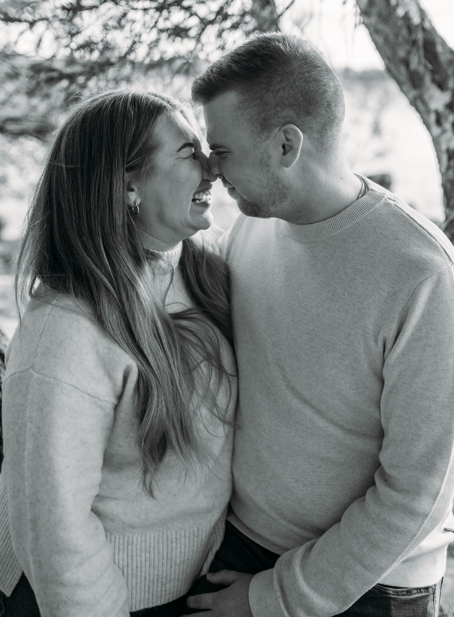 Black and white photo of a smiling couple touching noses affectionately outdoors near trees.