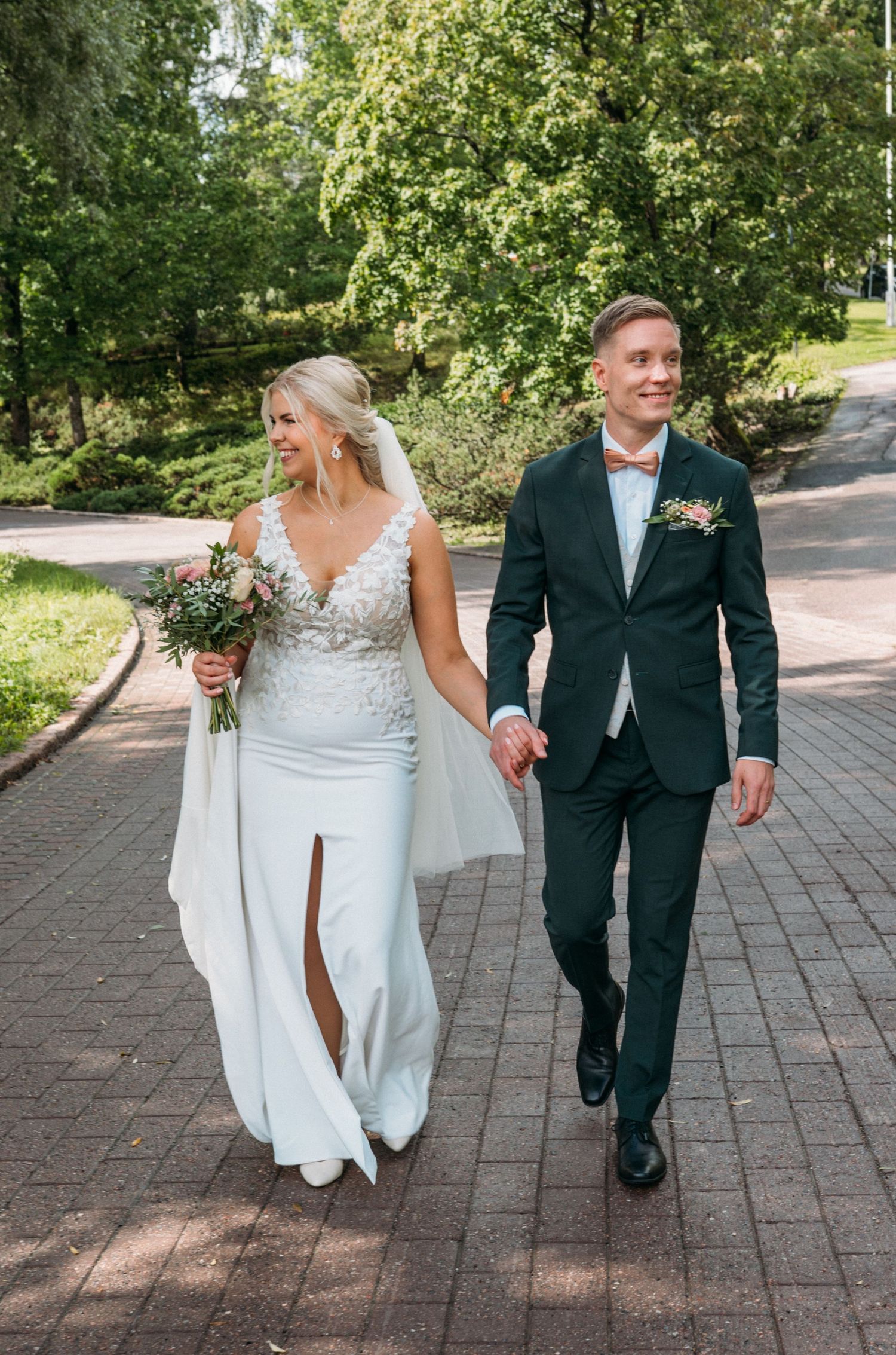 Happy bride and groom holding hands walking along a sunny tree-lined brick pathway.