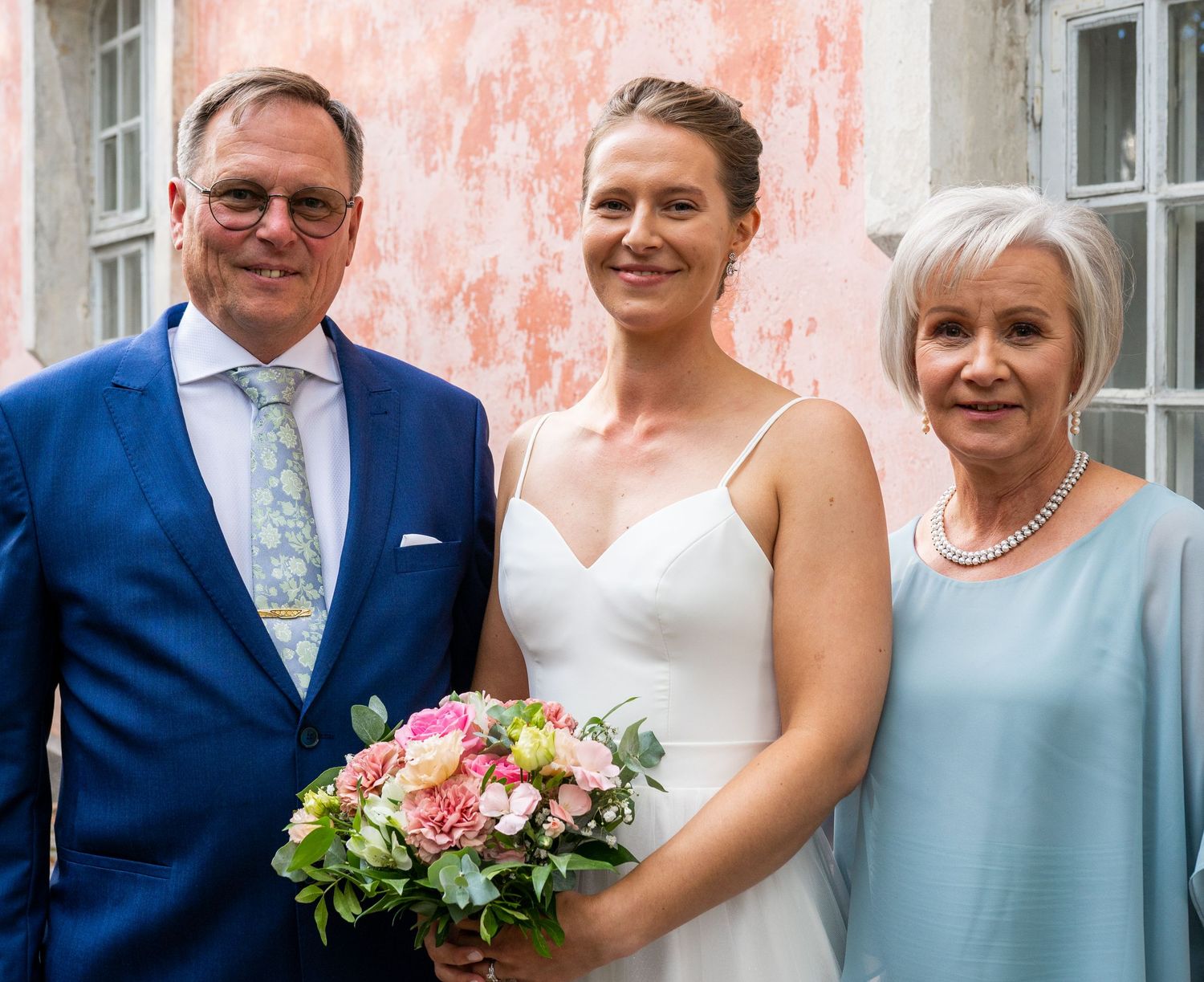Bride in white gown holds pink bouquet flanked by parents in blue suit and light blue dress before pink wall.