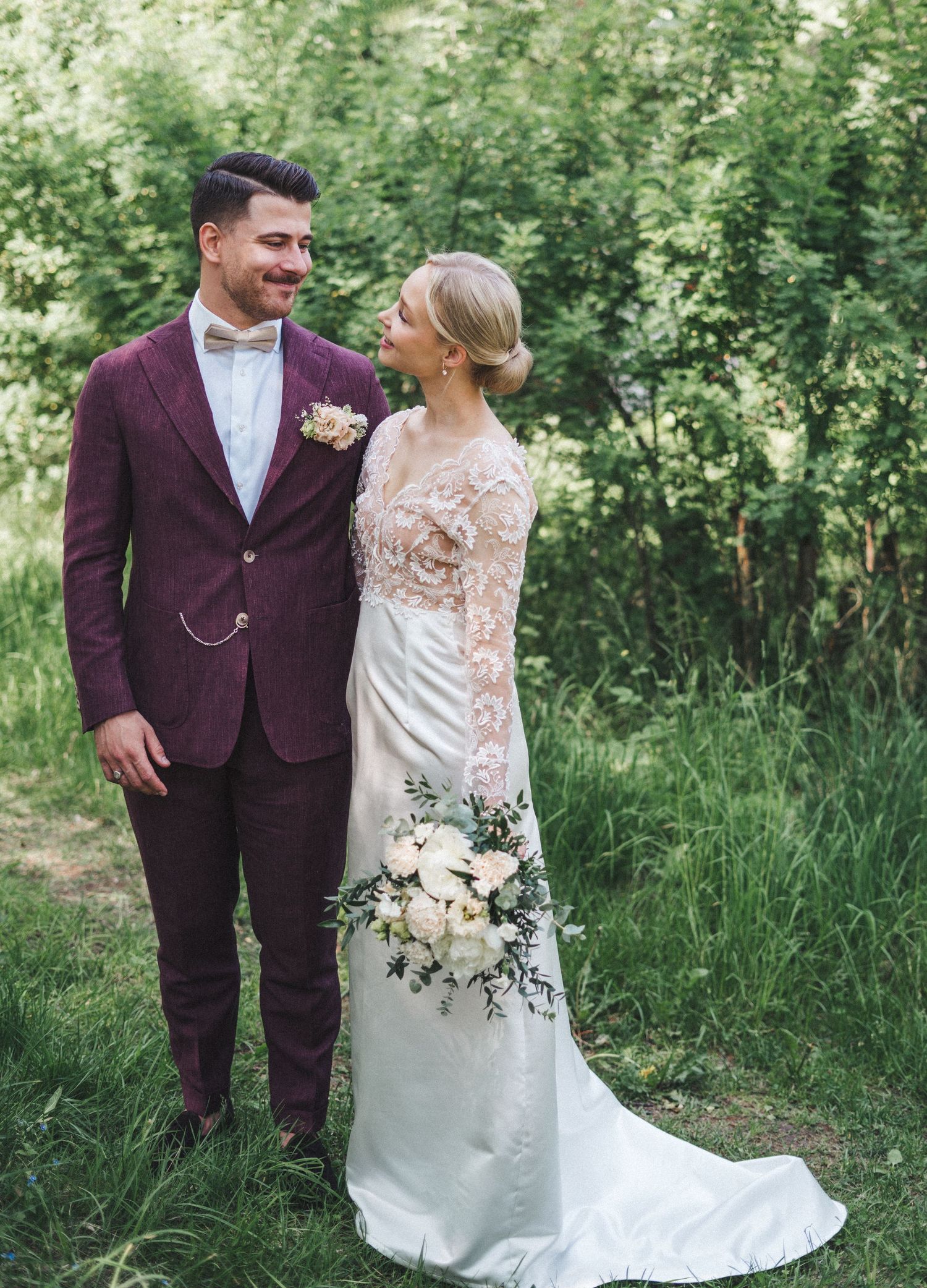 Wedding couple standing in green garden, groom in burgundy suit smiling at blonde bride in lace gown.