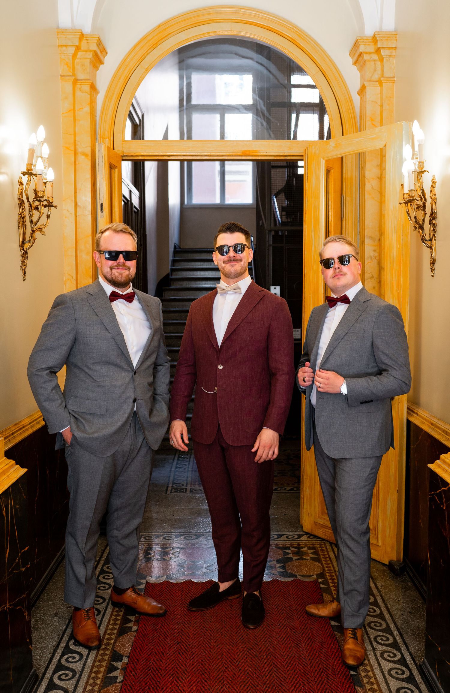 Three groomsmen in grey suits and burgundy bow ties posing in an elegant hallway archway.